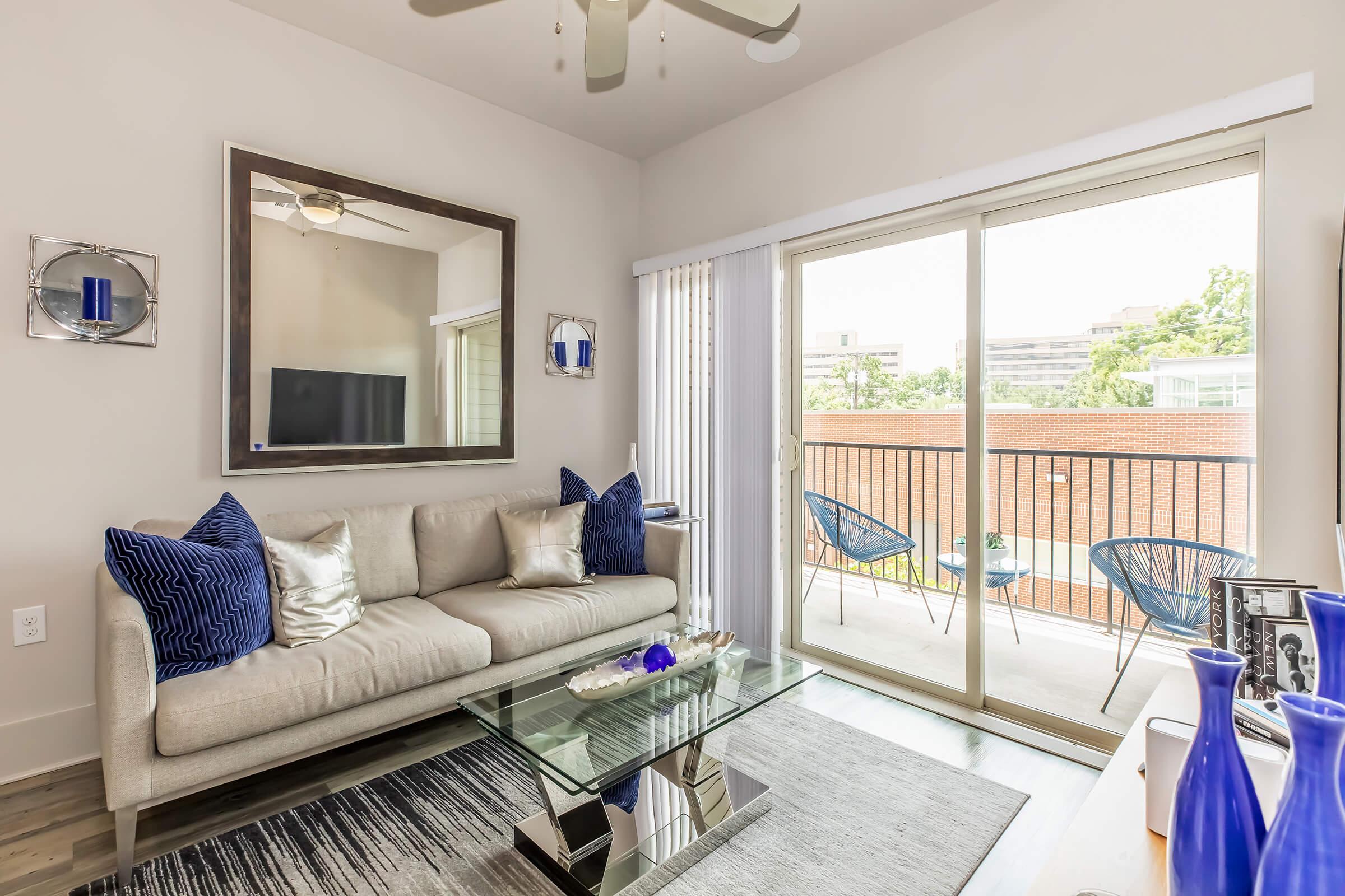 A modern living room featuring a light gray sofa with blue accent pillows, a glass coffee table on a striped rug, and large windows that open to a balcony. Decorative items in blue and white are placed on the table, enhancing the contemporary decor. A mirrored wall adds depth to the space.