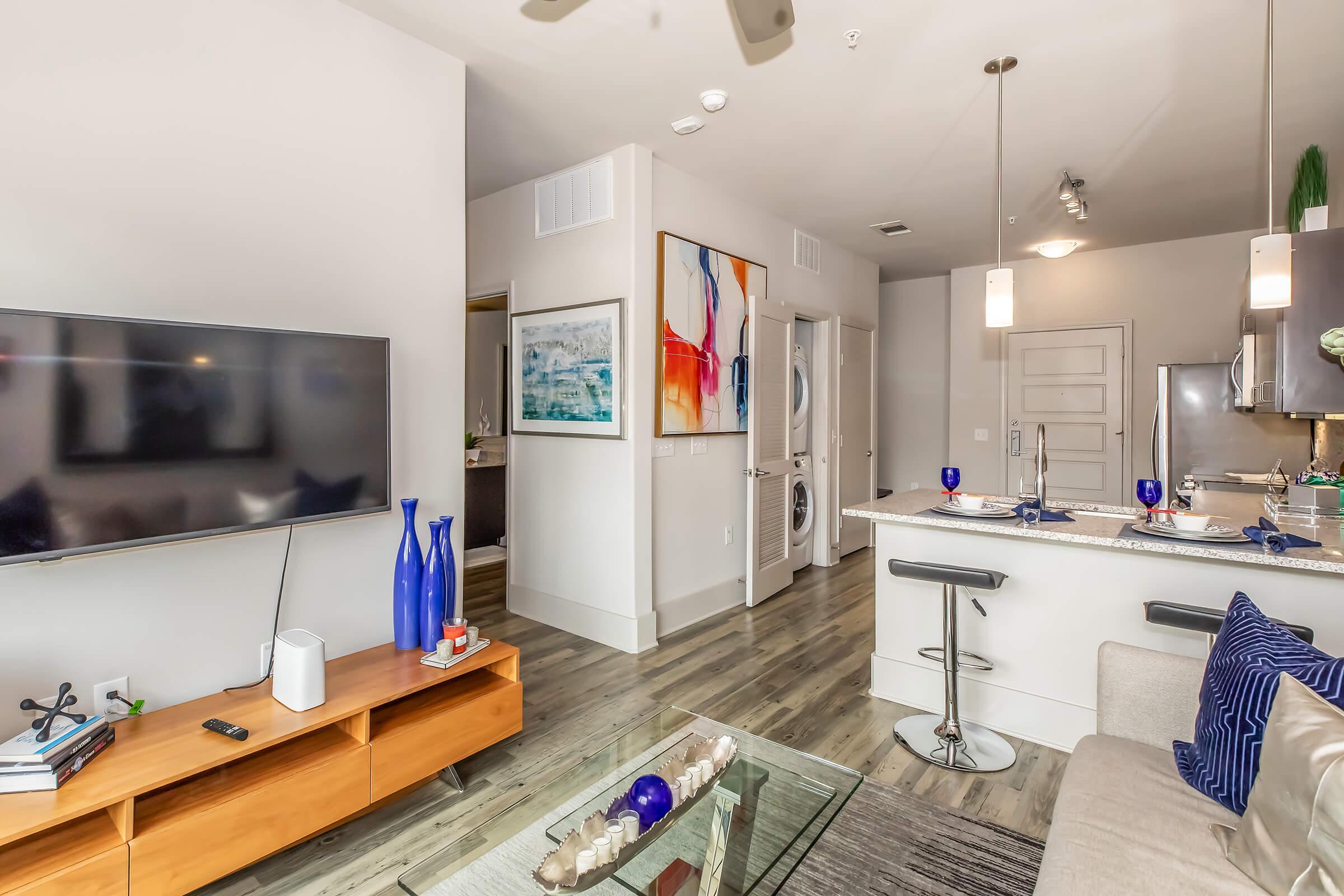 A modern living space featuring a stylish open-plan design. The room includes a television mounted on a light-colored wall, a cozy sofa, a glass coffee table adorned with decorative items, and a kitchen area with bar stools. Bright artwork hangs on the wall, and there are subtle decorative elements in blue tones.