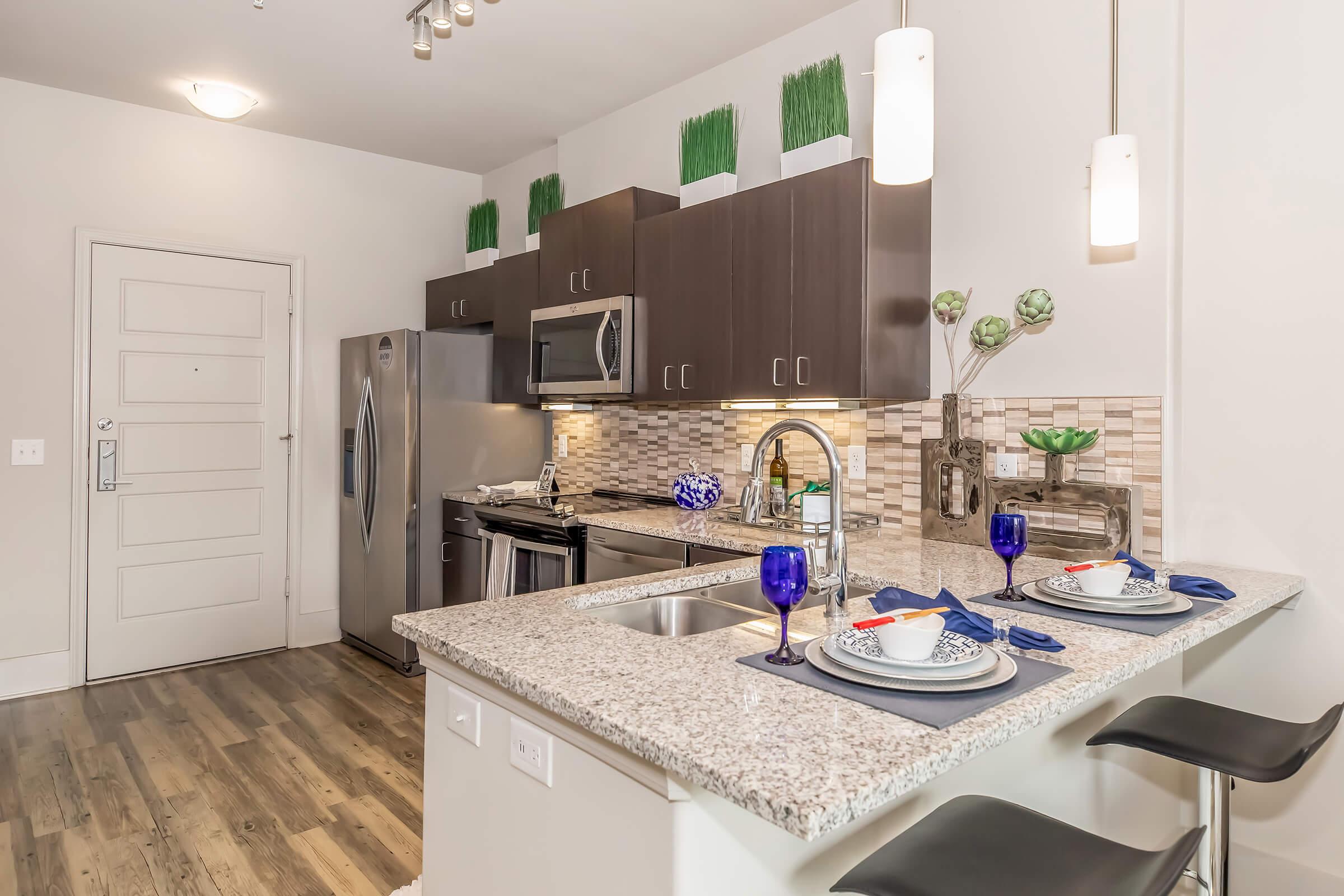 A modern kitchen featuring dark cabinetry, stainless steel appliances, and a granite countertop. Two place settings with blue glasses and plates are set on the island, which has bar stools. The backsplash consists of mosaic tiles, and green plants are displayed above the cabinets. Bright lighting enhances the space.