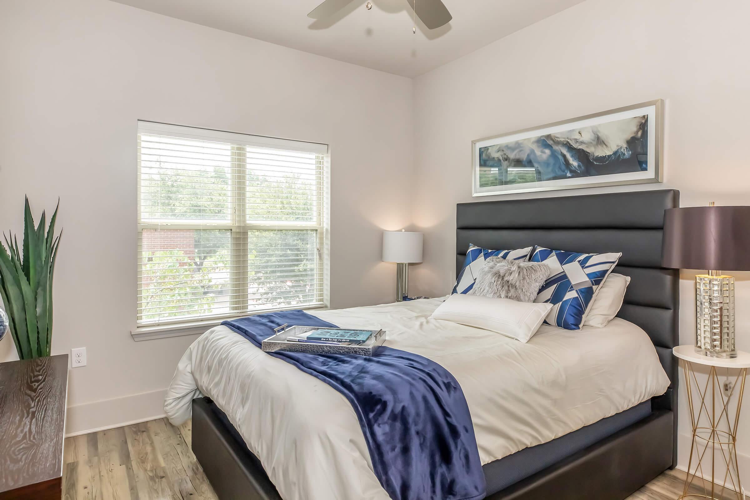 A cozy bedroom featuring a modern design with a comfortable bed adorned with blue and white pillows and a plush throw. There is a stylish lamp on a bedside table, a large window with natural light, and a decorative plant in the corner, creating a welcoming atmosphere.