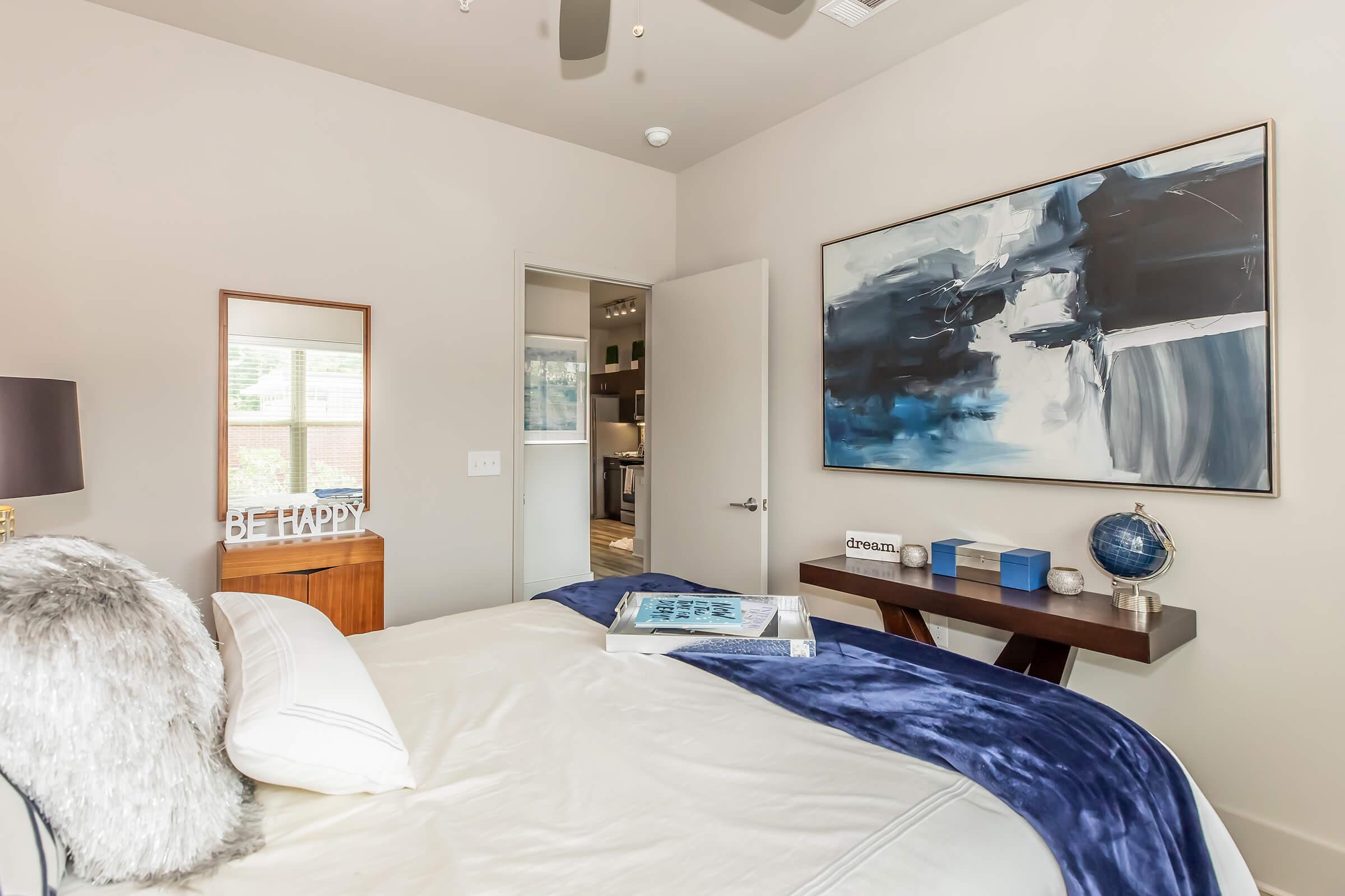 A modern bedroom featuring a bed with a white comforter and navy blue throw. There's a decorative mirror and a shelf with a globe and decorative items. A large abstract painting in shades of blue hangs on the wall, and a door leads to another room. The overall decor is minimalist and stylish.