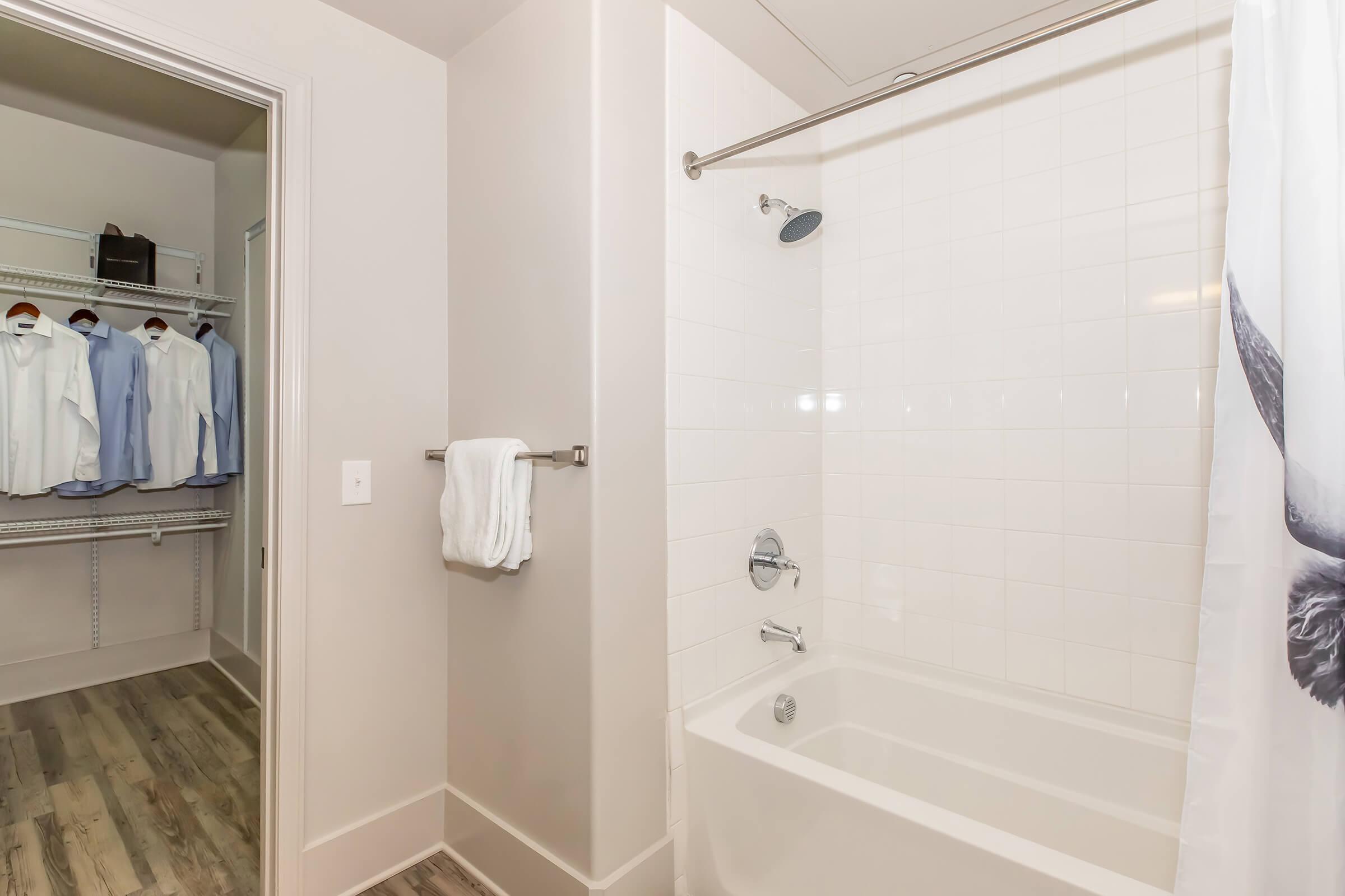 A clean bathroom featuring a bathtub with a shower head and a glass shower curtain. On the left, a walk-in closet with a rack displaying neatly hung light-colored shirts. Modern tiled flooring complements the overall aesthetic.