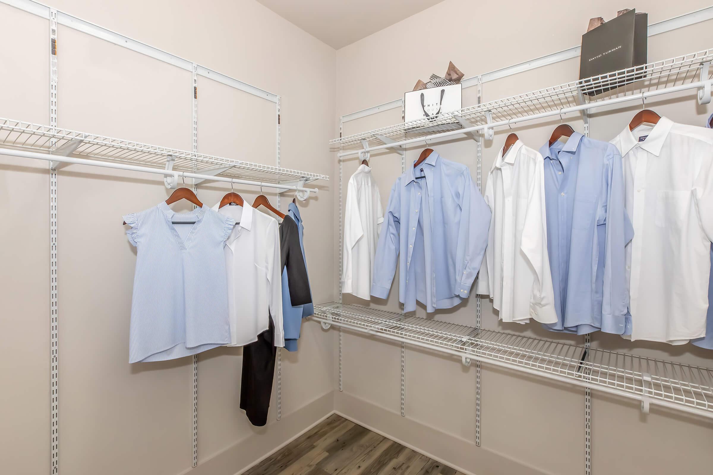 A well-organized walk-in closet featuring white and light blue dress shirts hanging on wire shelves. One pair of black pants hangs alongside the shirts, and a box is placed on a shelf above. The walls are neutral, and the flooring is light wood.