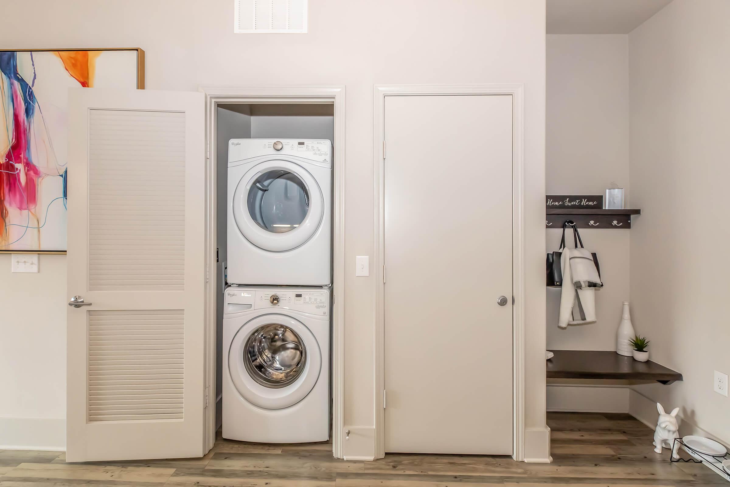 A compact laundry area featuring a stacked washer and dryer in a closet, flanked by two doors. To the right, there's a small entryway with a shelf for storage, hooks for hanging items, and a decorative plant, all set against a light-colored wall and wooden floor.