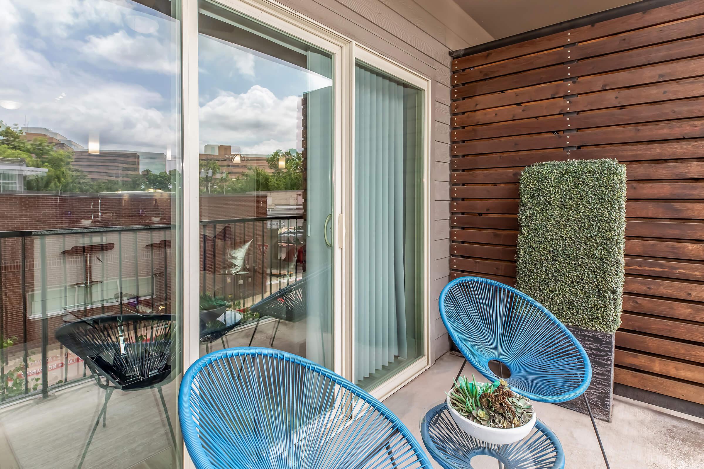 A modern balcony featuring two bright blue chairs and a potted plant, with a large decorative planter covered in greenery. The balcony is enclosed by a glass sliding door and has a wooden accent wall, creating a stylish outdoor space with a view of nearby buildings and a cloudy sky.
