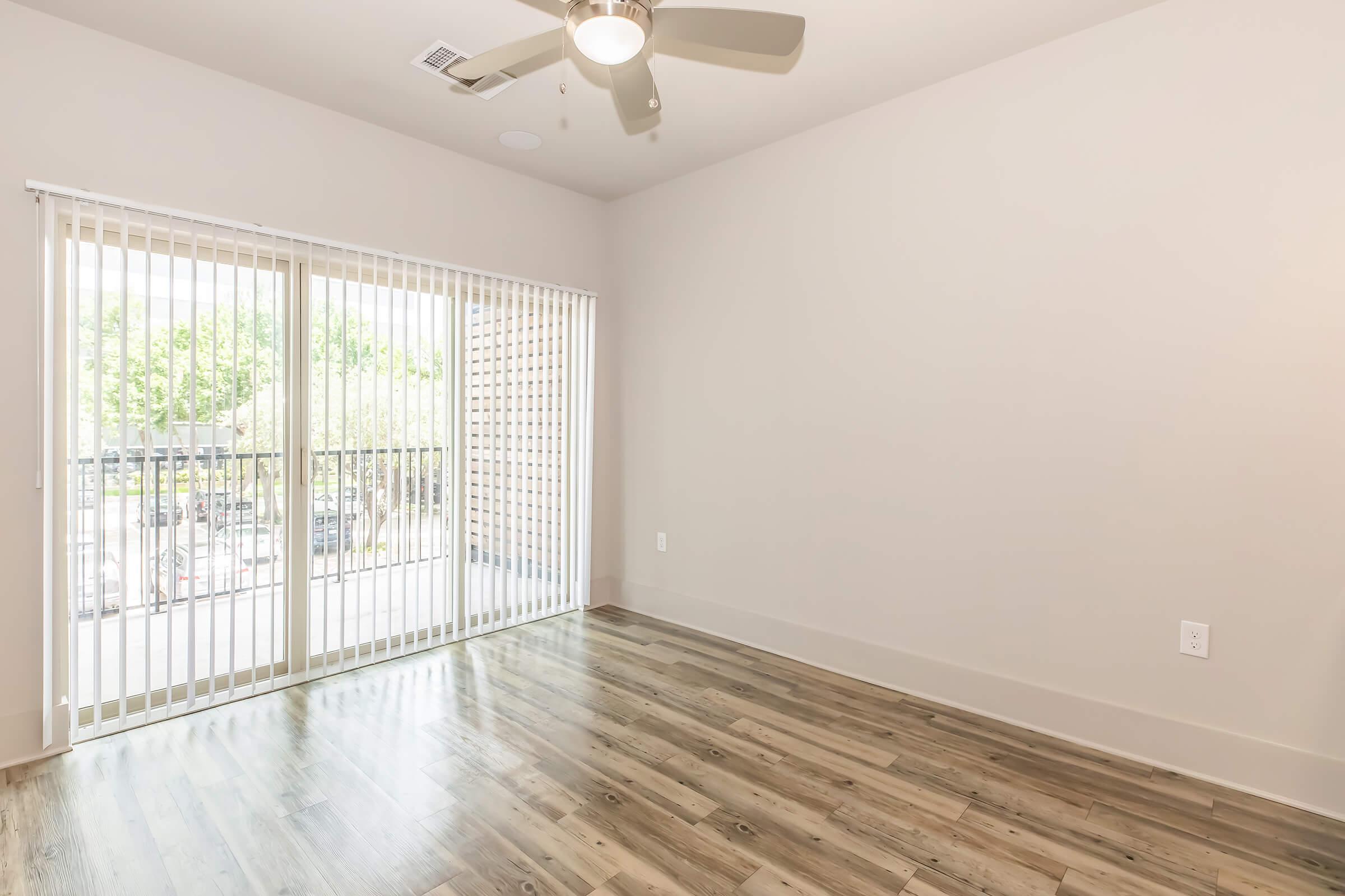 A spacious, empty room with light-colored walls and a ceiling fan. Large sliding glass doors with vertical blinds lead to a balcony overlooking an outdoor area. The floor is made of light hardwood or laminate. Natural light floods the space, creating a bright atmosphere.