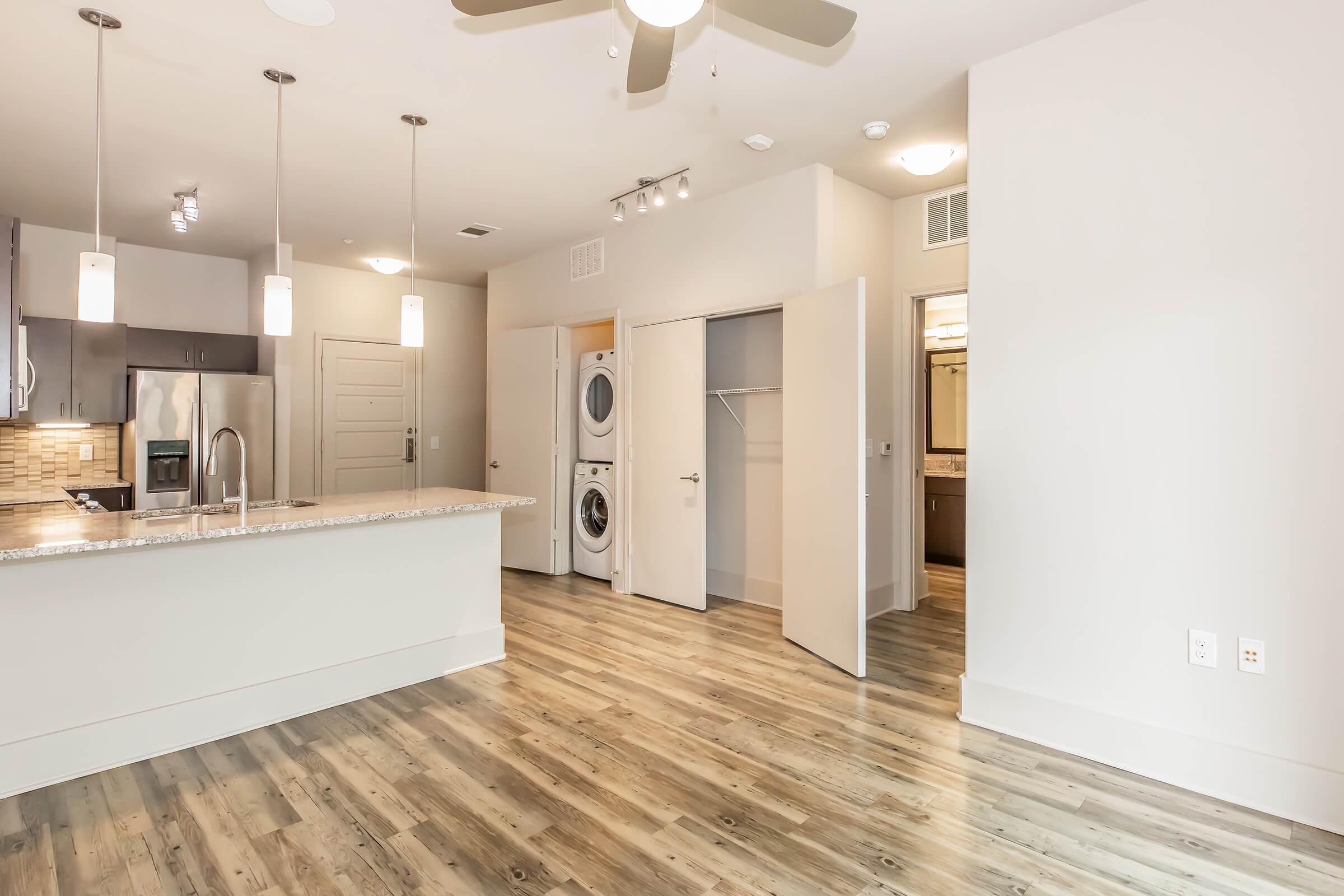 Interior view of a modern apartment showcasing an open layout. Features include a kitchen with a granite countertop, stainless steel appliances, a ceiling fan, and wooden flooring. Visible are a laundry area with stacked washer and dryer, a bathroom entrance, and a closet. The walls are painted in neutral tones.
