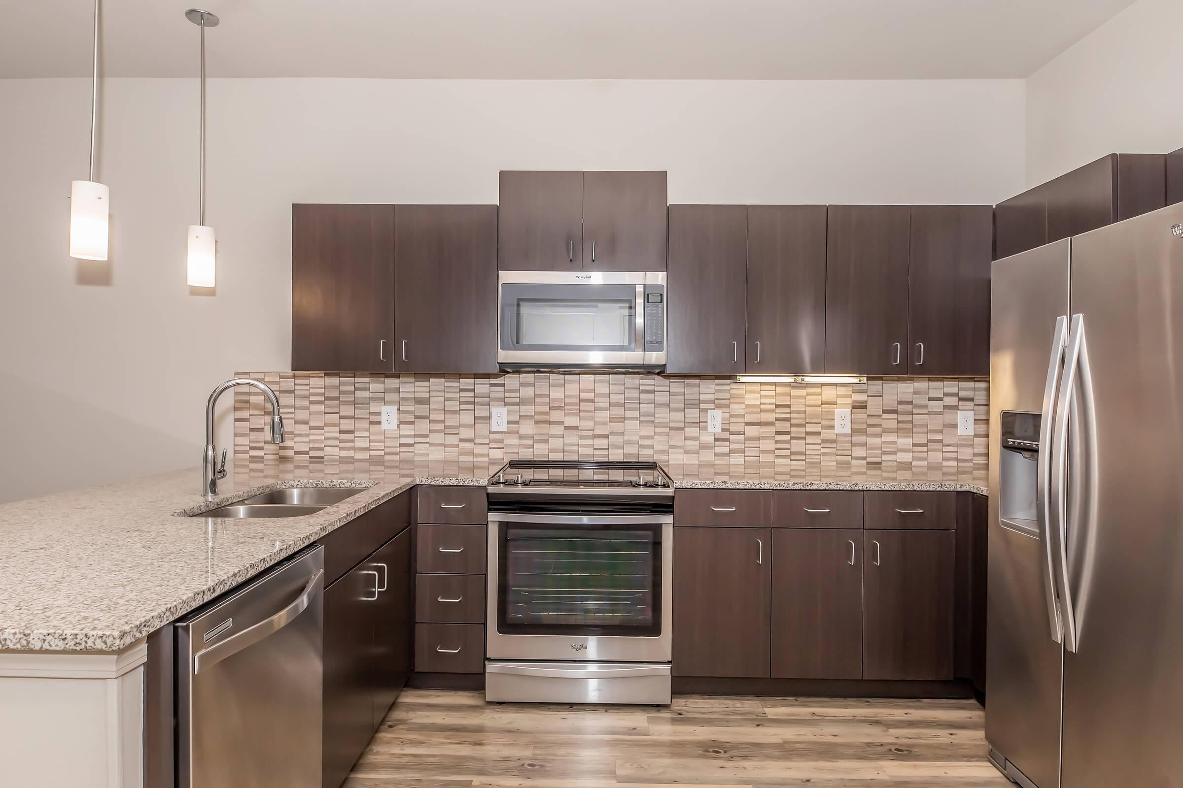 Modern kitchen featuring dark wood cabinetry, stainless steel appliances including a refrigerator, oven, and microwave, granite countertops, and a tiled backsplash. The space is well-lit with pendant lights and has a contemporary, clean design.