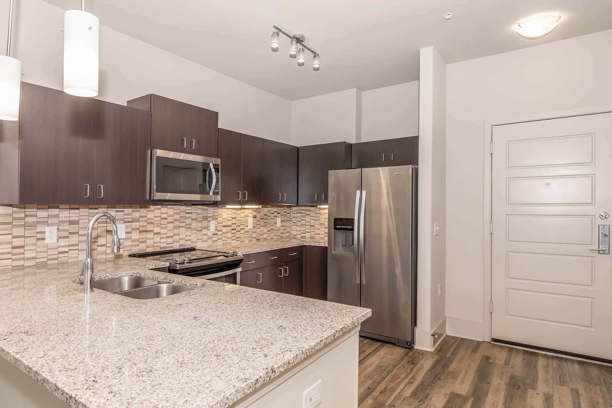 Modern kitchen featuring dark wood cabinets, stainless steel appliances, and a granite countertop. The backsplash is made of tiled mosaics, complemented by under-cabinet lighting. A stainless steel refrigerator is visible alongside an oven and microwave, with a spacious entrance area to the side.