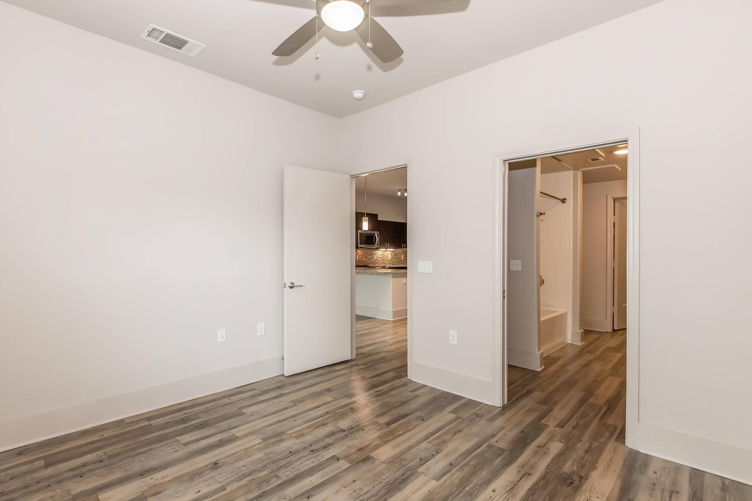 A spacious, well-lit room featuring light-colored walls and wood-like flooring. An open doorway leads to a bathroom, while another door opens to a kitchen area visible in the background. A ceiling fan is installed, adding to the modern aesthetic of the interior.