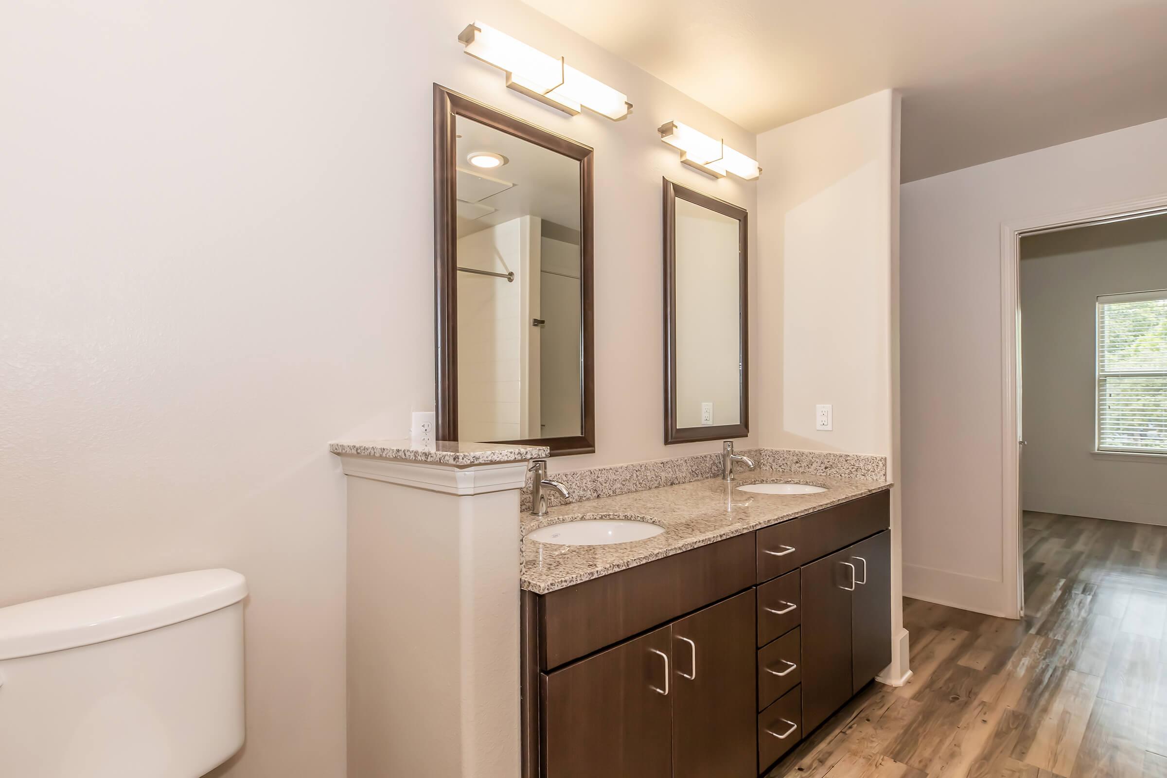 A modern bathroom featuring a double vanity with two sinks, granite countertops, and two mirrors above. There is a toilet to the left, and the floor is finished with wood-look tiles. A window can be seen in the background, allowing natural light to brighten the space.