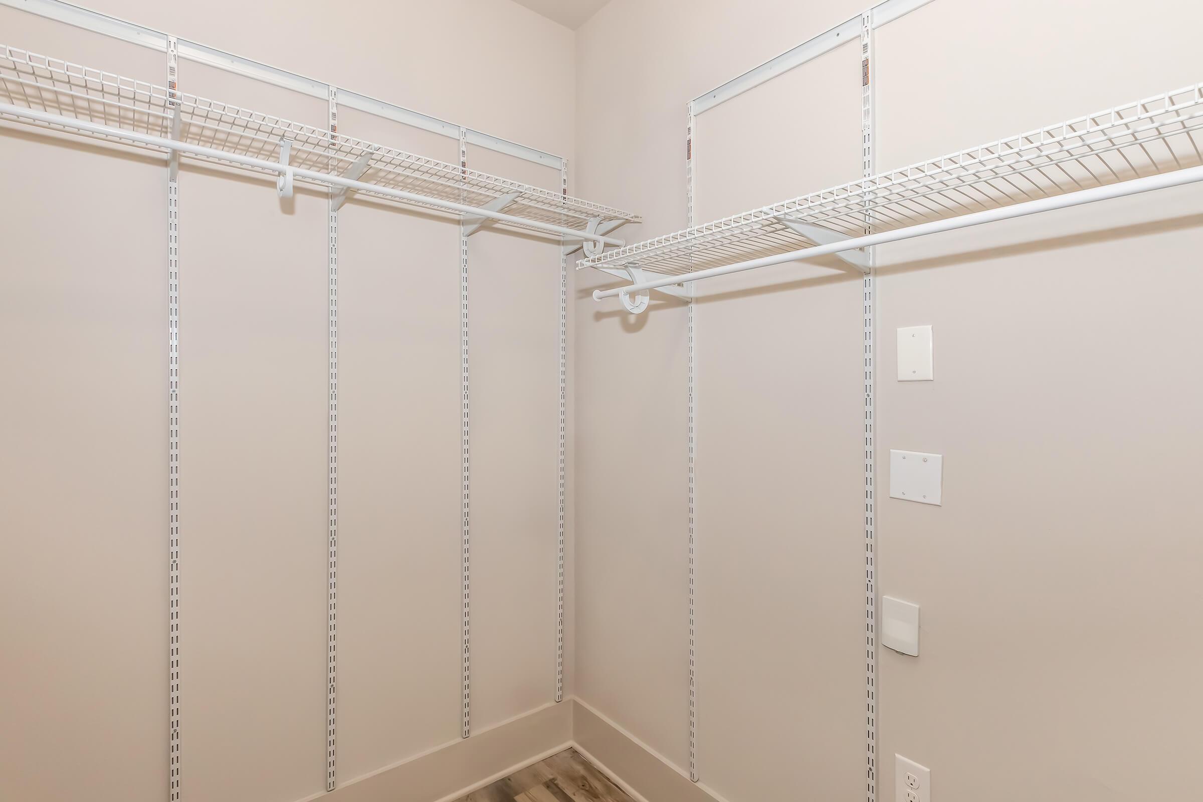 A spacious empty closet with wire shelving and a neutral-colored wall. The closet features two hanging shelves on either side, with no items stored. The flooring is light-colored wood, enhancing the bright and open feel of the space.