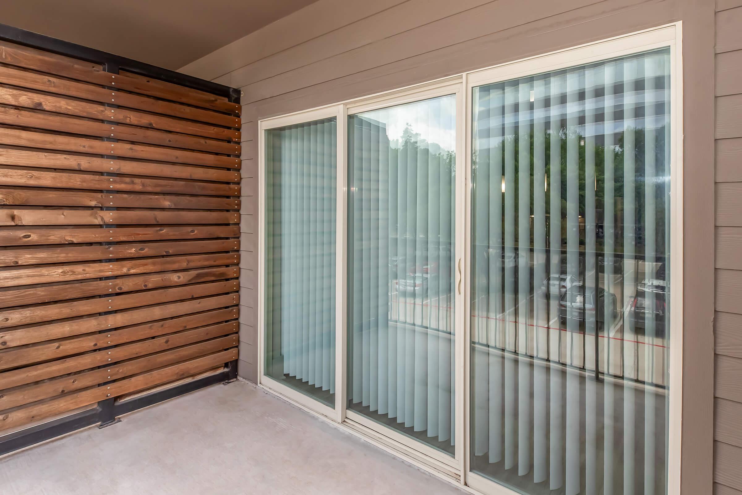 A modern balcony featuring sliding glass doors with vertical blinds and a wooden accent wall on one side. The flooring is concrete, and there is a view of parked cars outside through the glass.