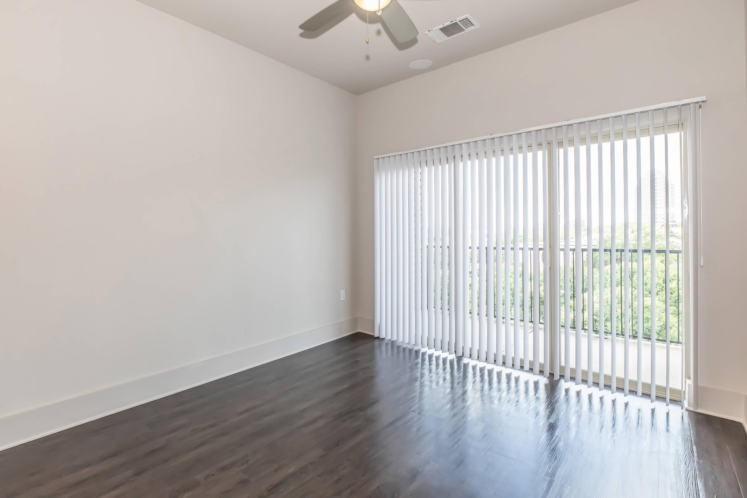 Bright, empty room with light-colored walls and dark wood flooring. A ceiling fan is visible, and large sliding glass doors with vertical blinds lead to a balcony or outdoor area. Natural light filters in, creating an open and airy atmosphere.