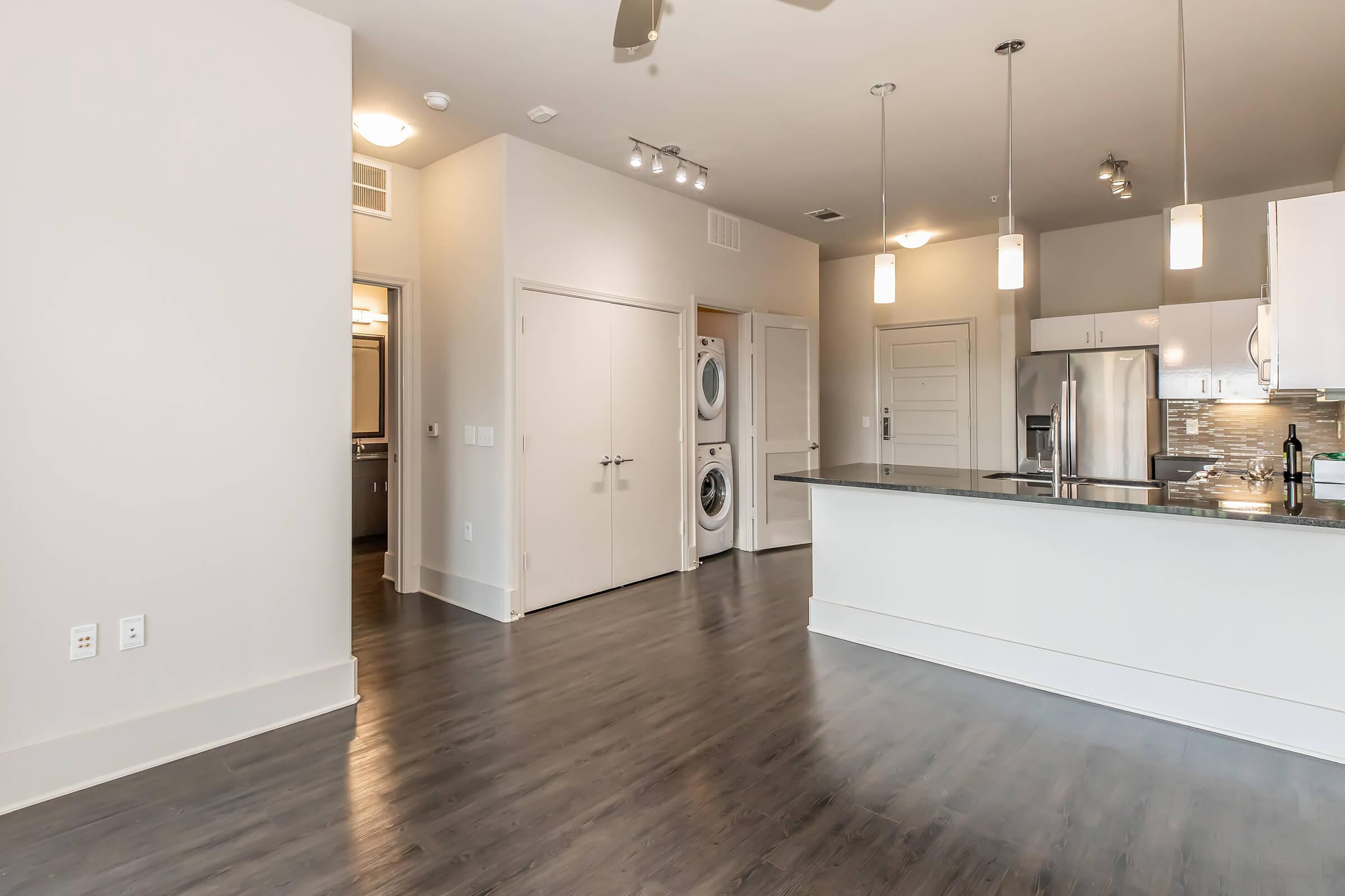 Modern open-concept apartment interior featuring a kitchen with stainless steel appliances, a black granite countertop, and a laundry area with stacked washer and dryer. The living space has light-colored walls and wide plank flooring, with recessed lighting and a ceiling fan enhancing the contemporary design.