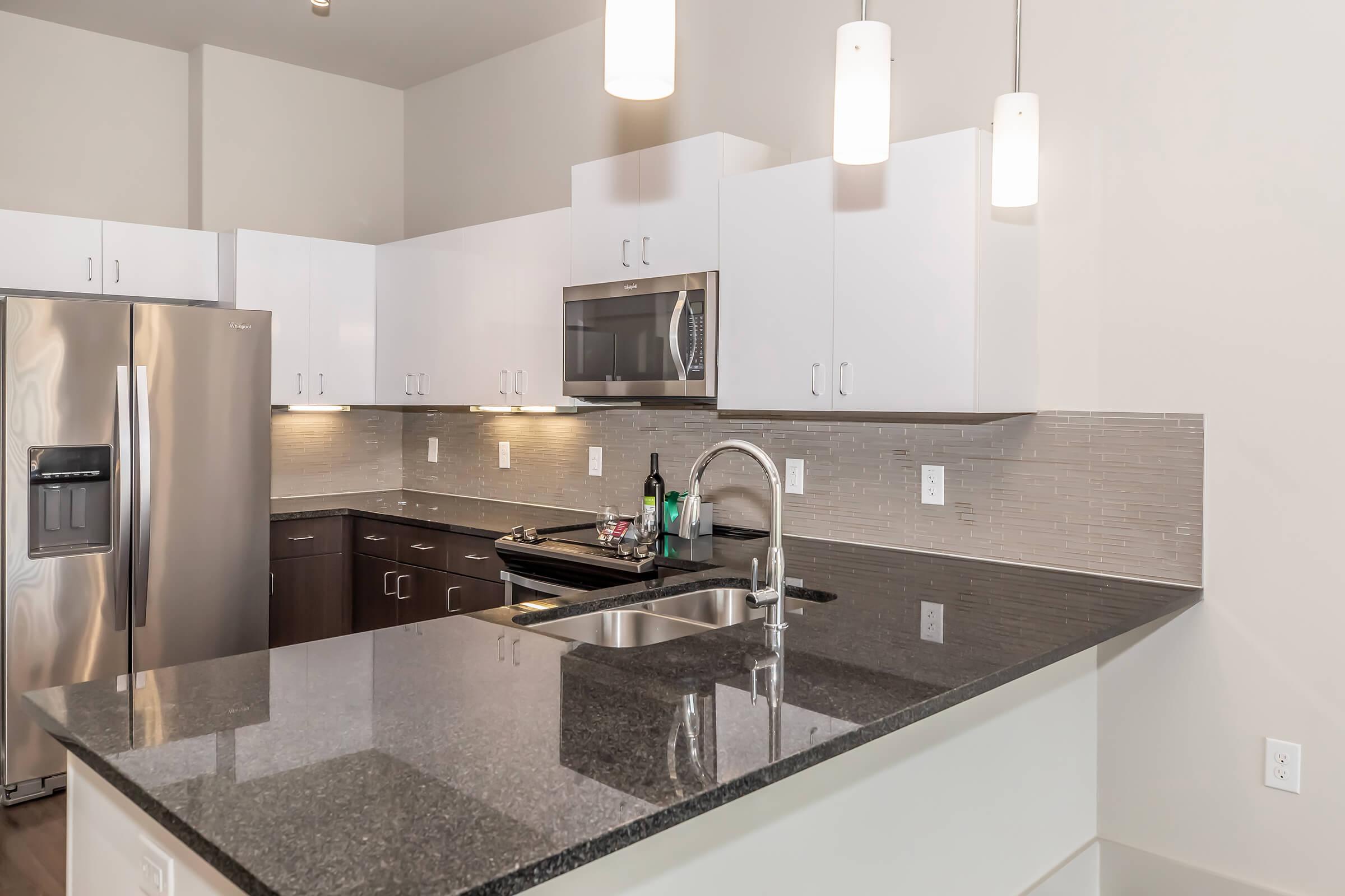 Modern kitchen featuring sleek white cabinets and a dark granite countertop. Stainless steel refrigerator and microwave are visible, along with a sink and a faucet. Pendant lights hang over the countertop, and neutral tones create a clean, contemporary look.