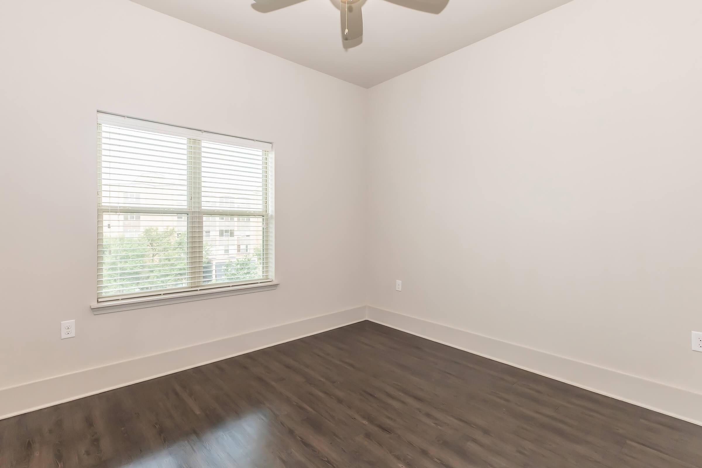 A bright, empty room featuring a ceiling fan and a large window with blinds. The walls are painted in a light color, and the floor is made of dark hardwood. Sunlight streams in through the window, illuminating the spacious area.