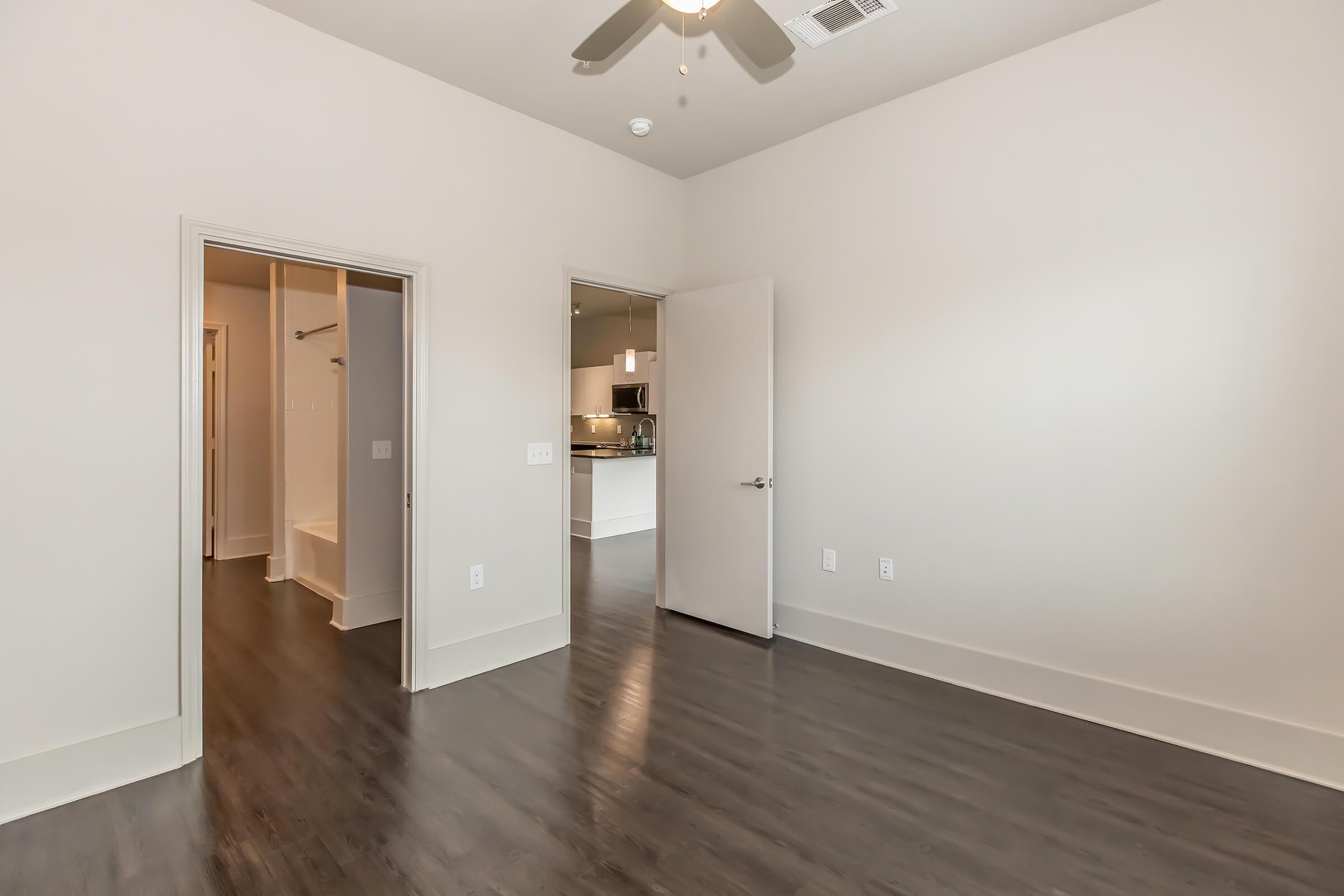 A spacious room with light-colored walls and dark wooden flooring. It features a ceiling fan and leads to a bathroom area on one side. An open doorway leads to a kitchen space in the background, visible through an adjacent door. The room is well-lit with natural light.