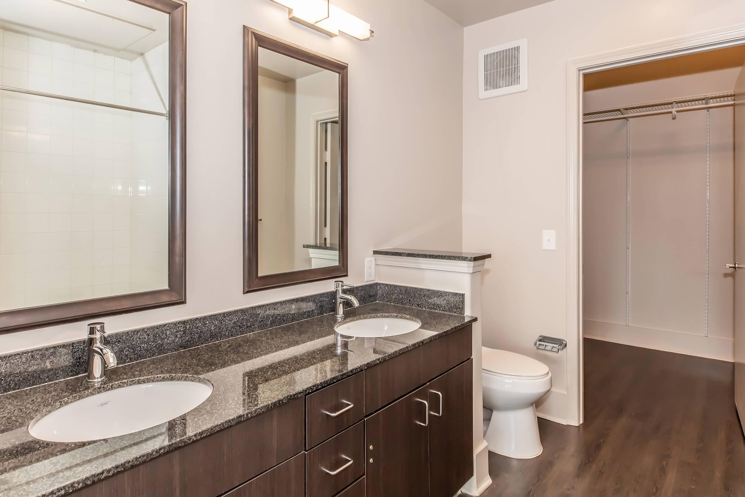 Brightly lit modern bathroom featuring dual sinks with rectangular mirrors above, dark wood cabinetry, and a granite countertop. A white toilet is positioned beside a spacious open closet with a sliding door in the background, showcasing a contemporary design with neutral wall colors.