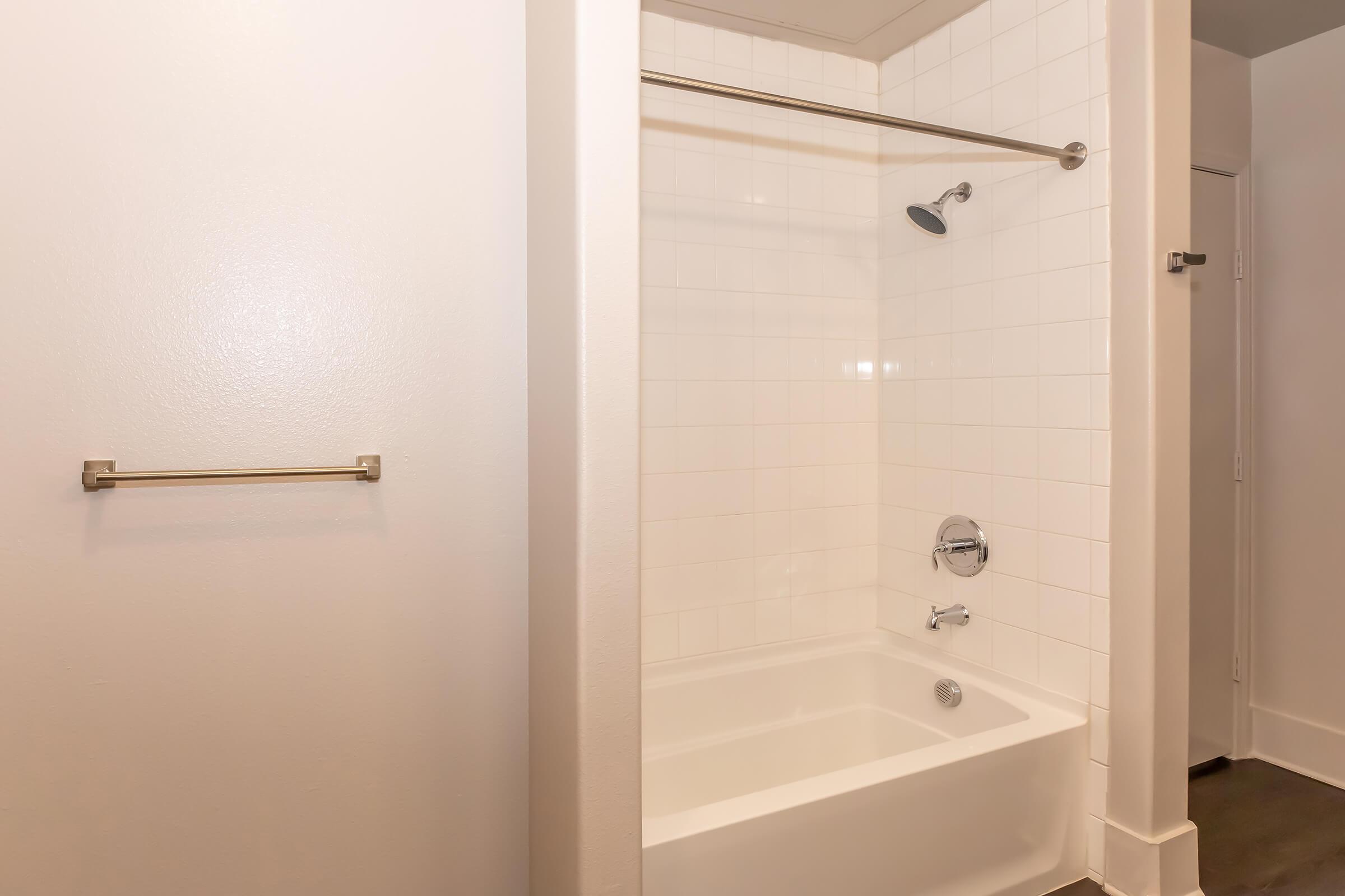 A clean, modern bathroom featuring a white tiled shower with a showerhead and a bathtub. The walls are painted in a light color, and there's a towel bar mounted on the wall. The floor is dark, contrasting with the lighter walls and tiles.