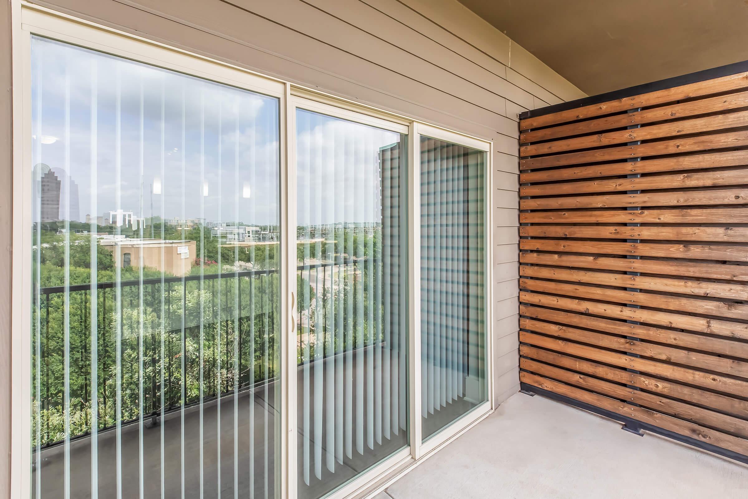 A modern balcony with sliding glass doors featuring vertical blinds. The balcony has a wooden privacy wall made of slatted wood panels. In the background, a view of city buildings and greenery can be seen under a partly cloudy sky.