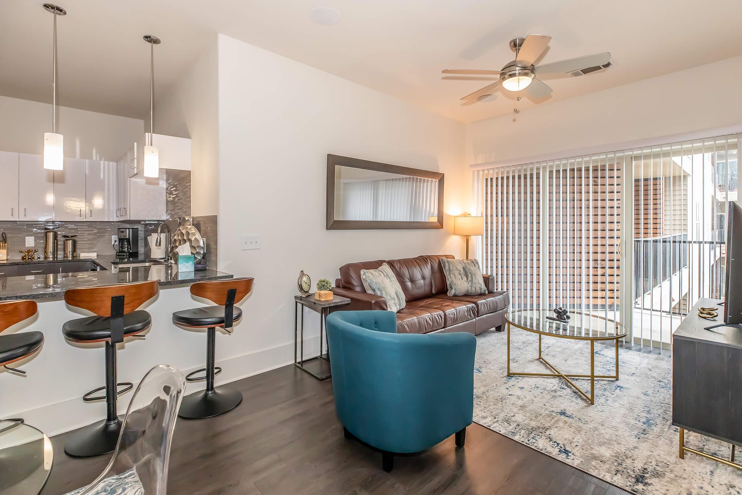 A modern living space featuring a brown leather sofa, a glass coffee table, and stylish blue accent chairs. A kitchen with bar stools is visible in the background, along with a large mirror and sliding glass doors that open to a balcony. The decor includes neutral tones with a cozy, contemporary feel.