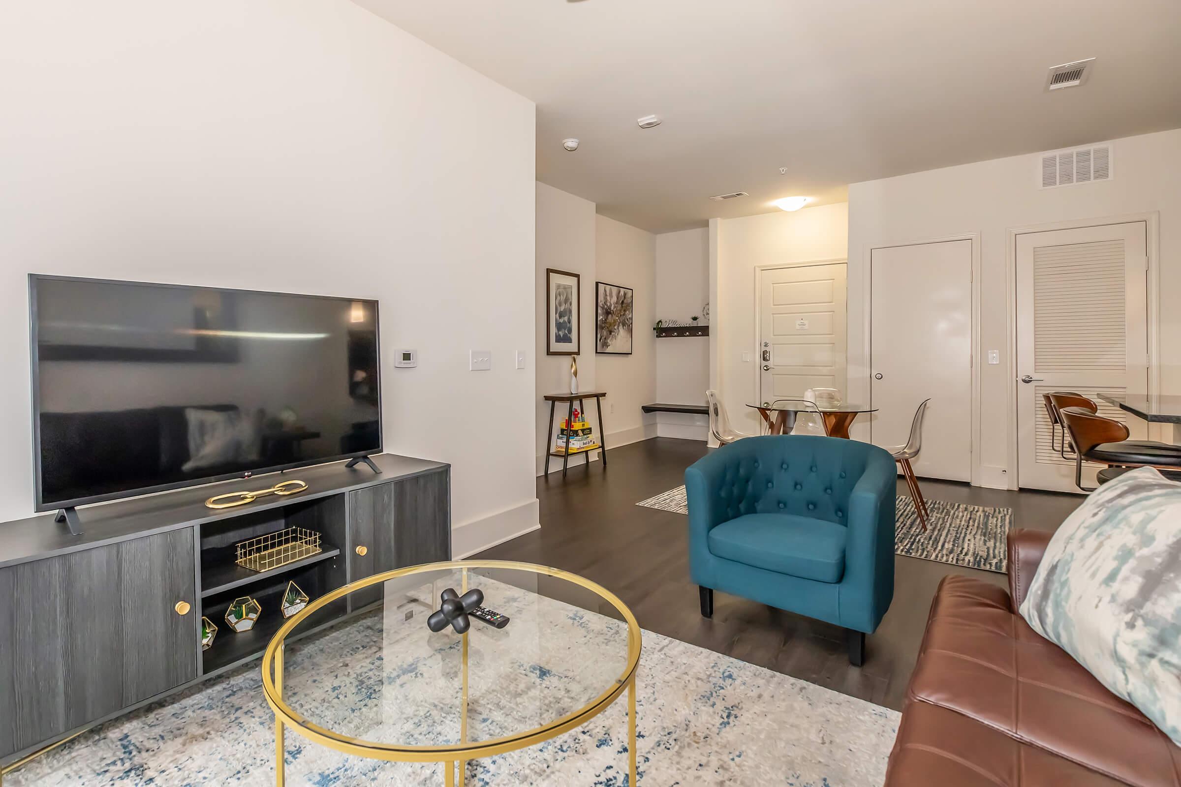 A modern living room featuring a flat-screen TV on a dark wooden console, a teal accent chair, a brown leather sofa, and a round dining table with clear chairs. There is a decorative rug on the floor and a doorway leading to another area, enhancing the contemporary aesthetic.