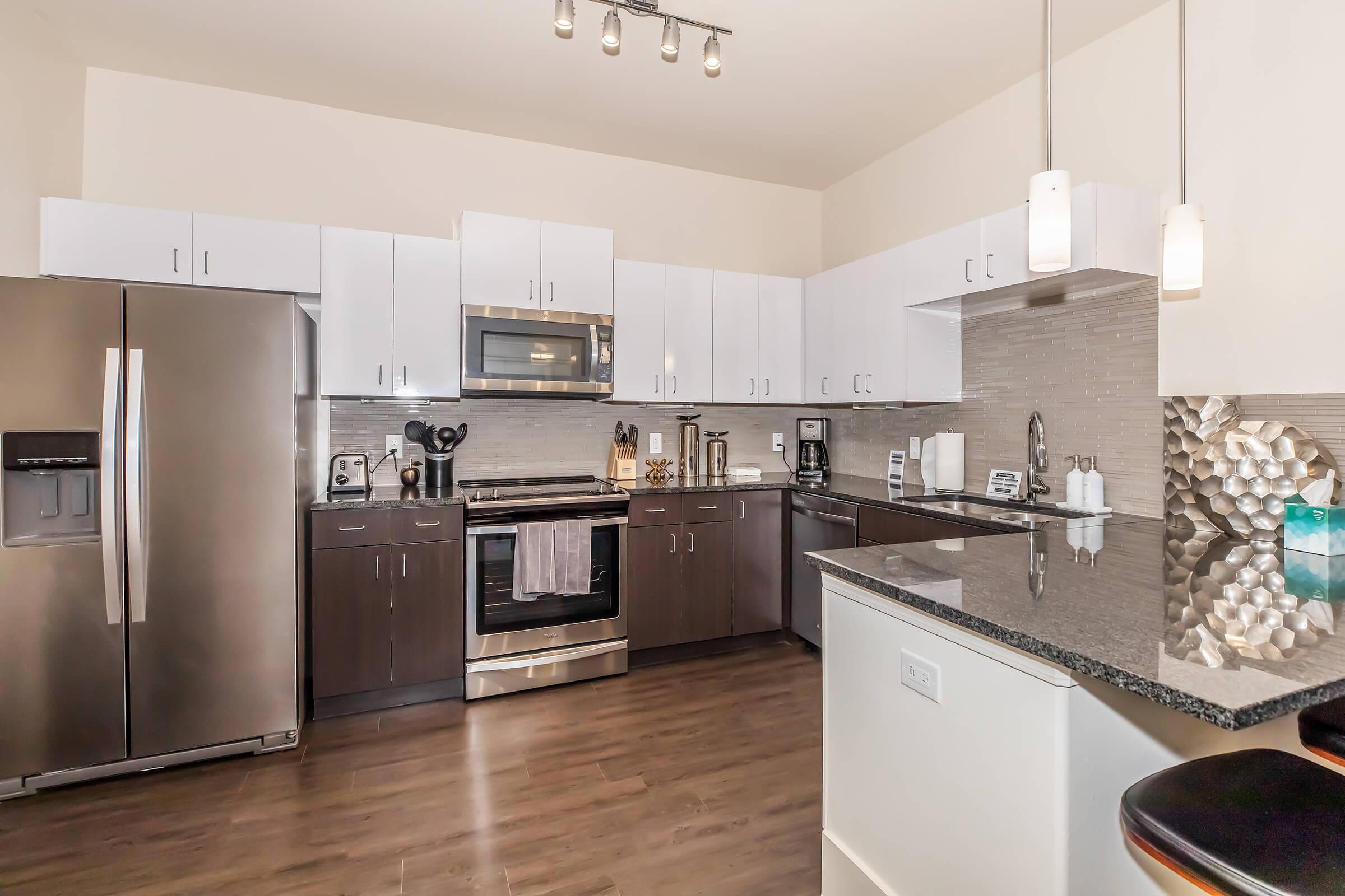 Modern kitchen featuring stainless steel appliances, including a refrigerator, oven, and microwave. Dark wood cabinets are paired with white upper cabinets, and a granite countertop. Stylish pendant lights hang above the counter, and ceramic mugs and a coffee maker are visible on the countertop.