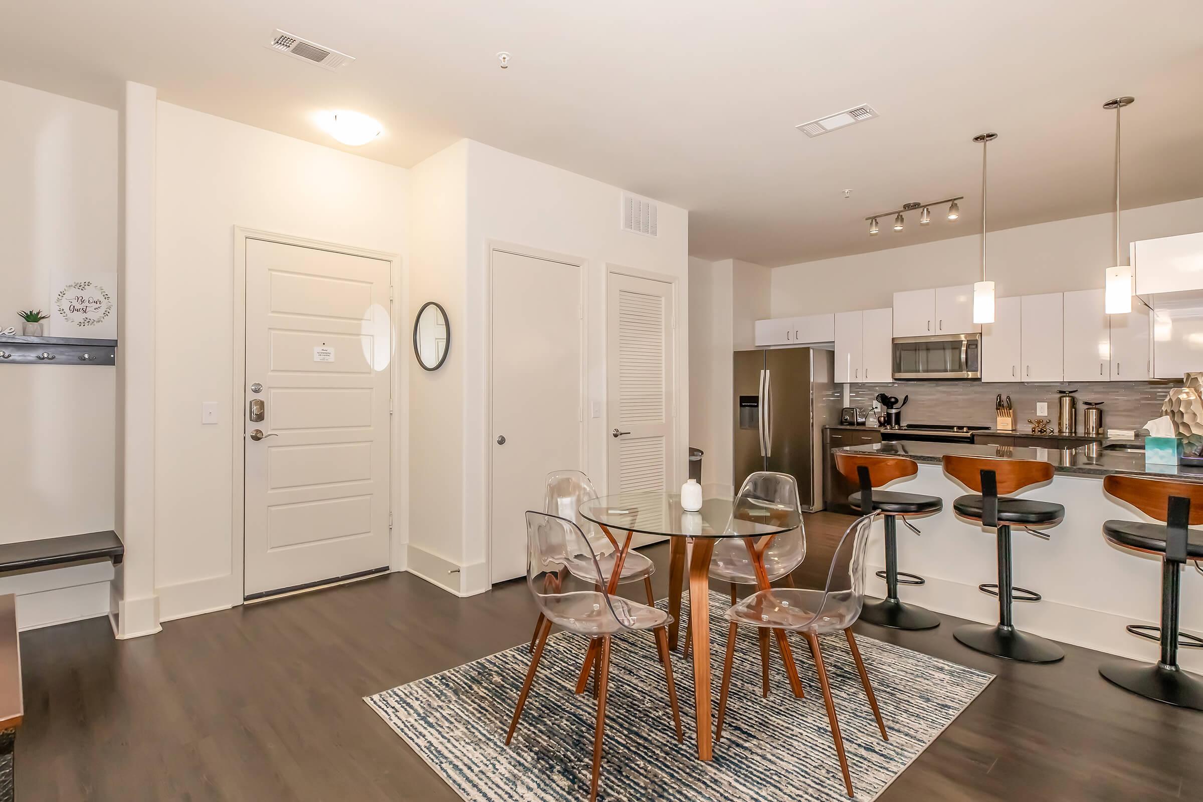 Modern apartment interior featuring an entryway with a round glass dining table and four clear chairs. In the background, a kitchen with stainless steel appliances and bar seating. Soft lighting from pendant lamps, hardwood floors, and a rug add warmth to the space.