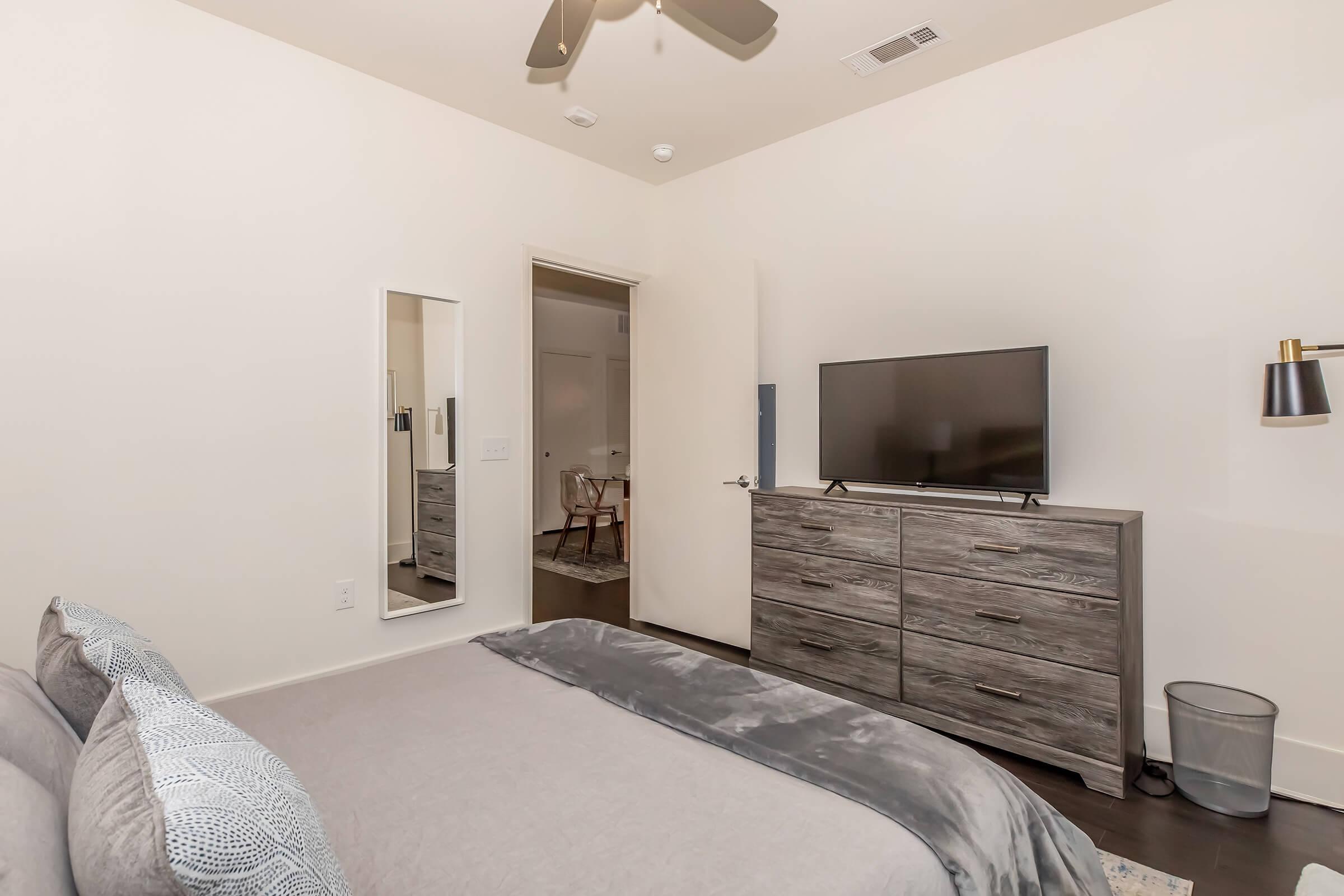 A modern bedroom featuring a bed with gray bedding, a wooden dresser with a flat-screen TV on top, and a mirror mounted on the wall. A light fixture with a shade is attached to the wall, and a door leads to another room. The flooring is dark, adding contrast to the light-colored walls.