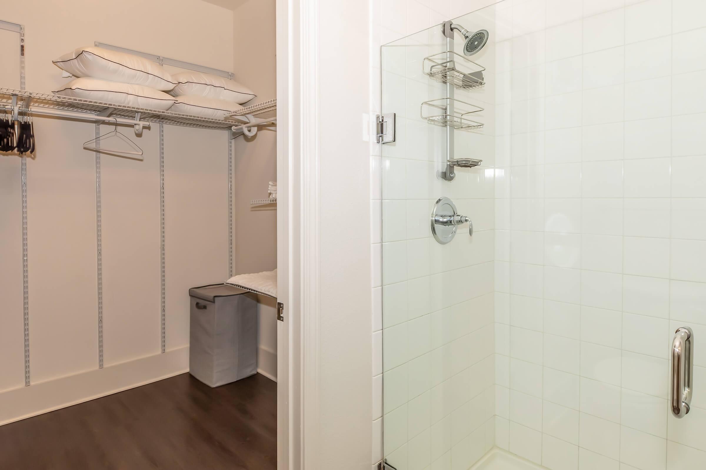 A clean bathroom featuring a glass shower enclosure and a minimalist design, alongside an organized closet space with shelves and hangers. Soft white pillows are neatly stored on a shelf, and a laundry bin is visible, creating a functional and modern look.