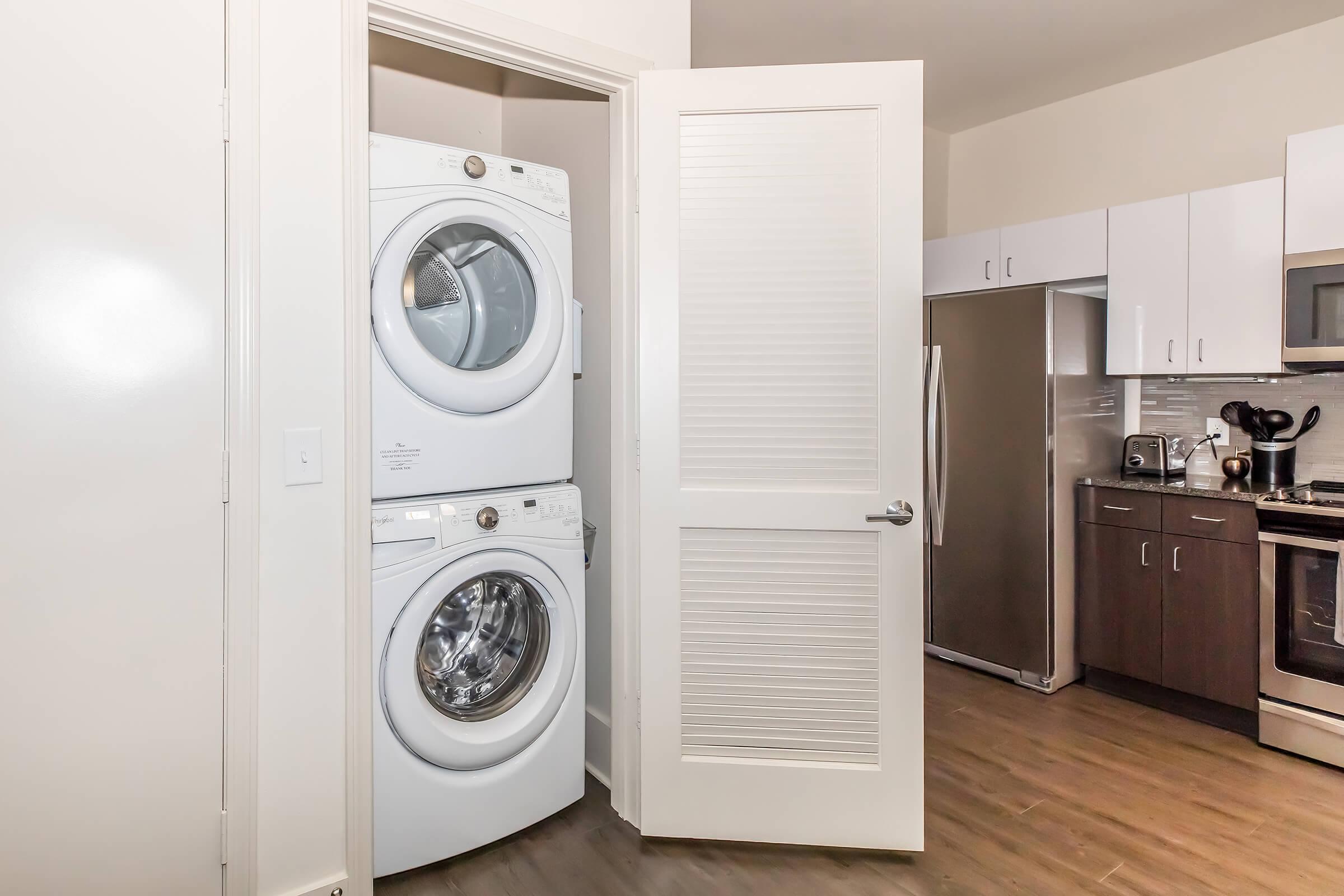 A laundry area featuring a stackable washer and dryer set positioned next to an open door. The room is part of a modern kitchen space with stainless steel appliances, including a refrigerator, and sleek cabinetry, all set against a light-colored wall and wooden flooring.