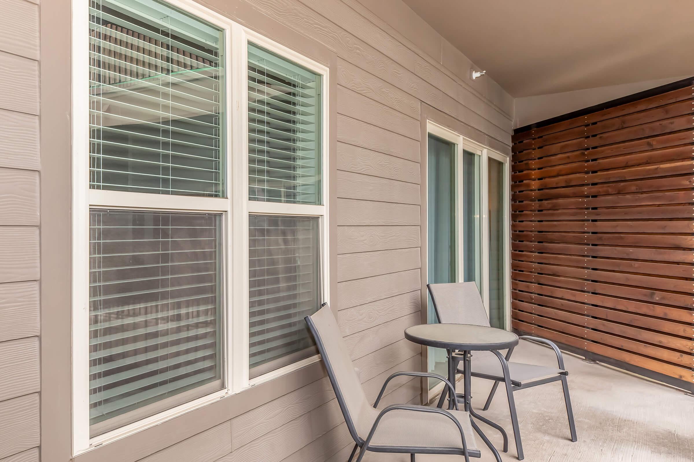 A small outdoor patio area featuring two metal chairs and a round table. The patio is bordered by a wooden fence and has large sliding glass doors that open to the interior. Sunlight highlights the light-colored walls with horizontal siding.