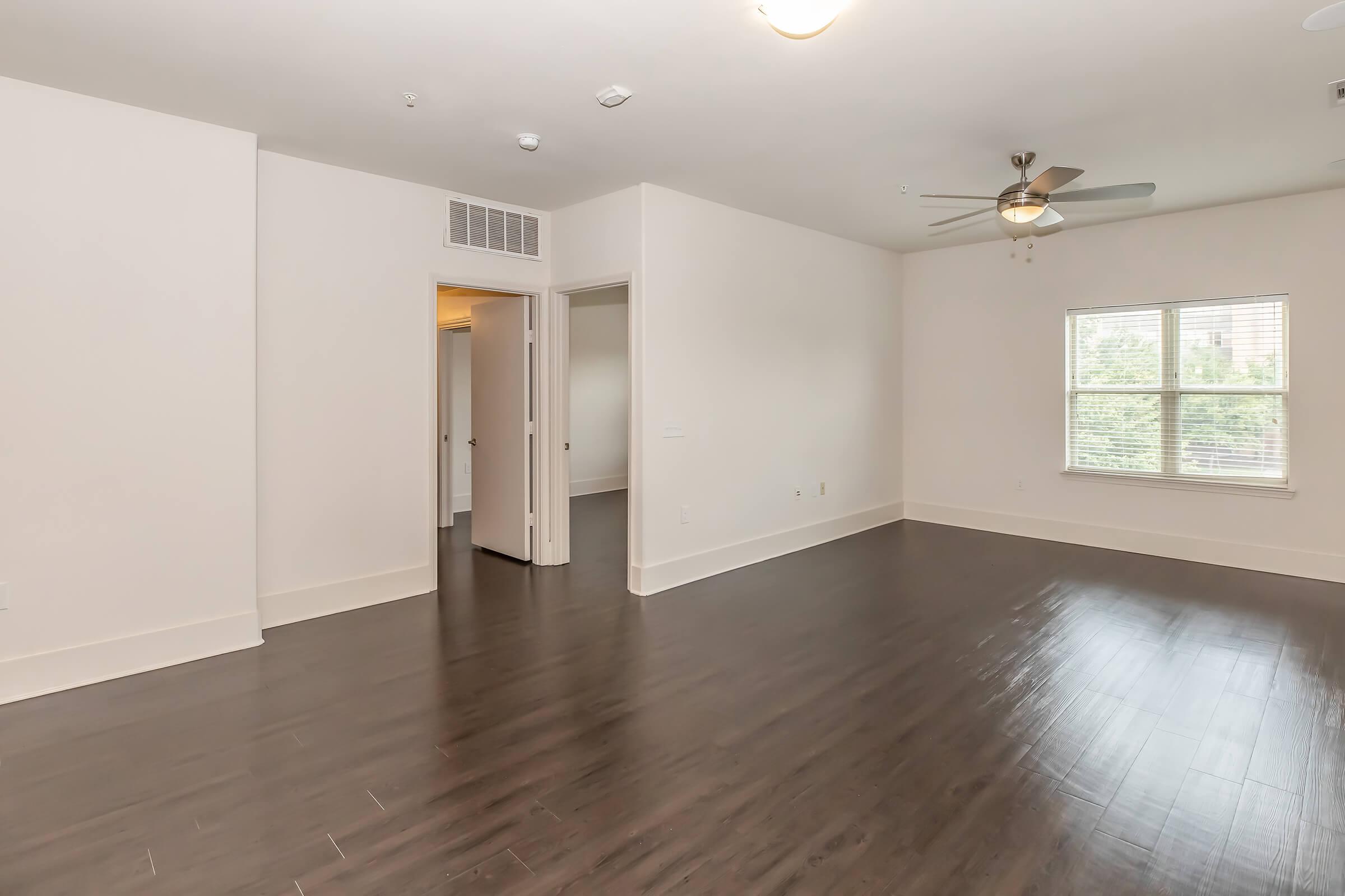 Spacious, modern living room showcasing dark hardwood floors, a ceiling fan, and a large window providing natural light. An open doorway leads to adjacent rooms, emphasizing the airy feel of the space. Walls are painted in a light color, enhancing the brightness of the area.