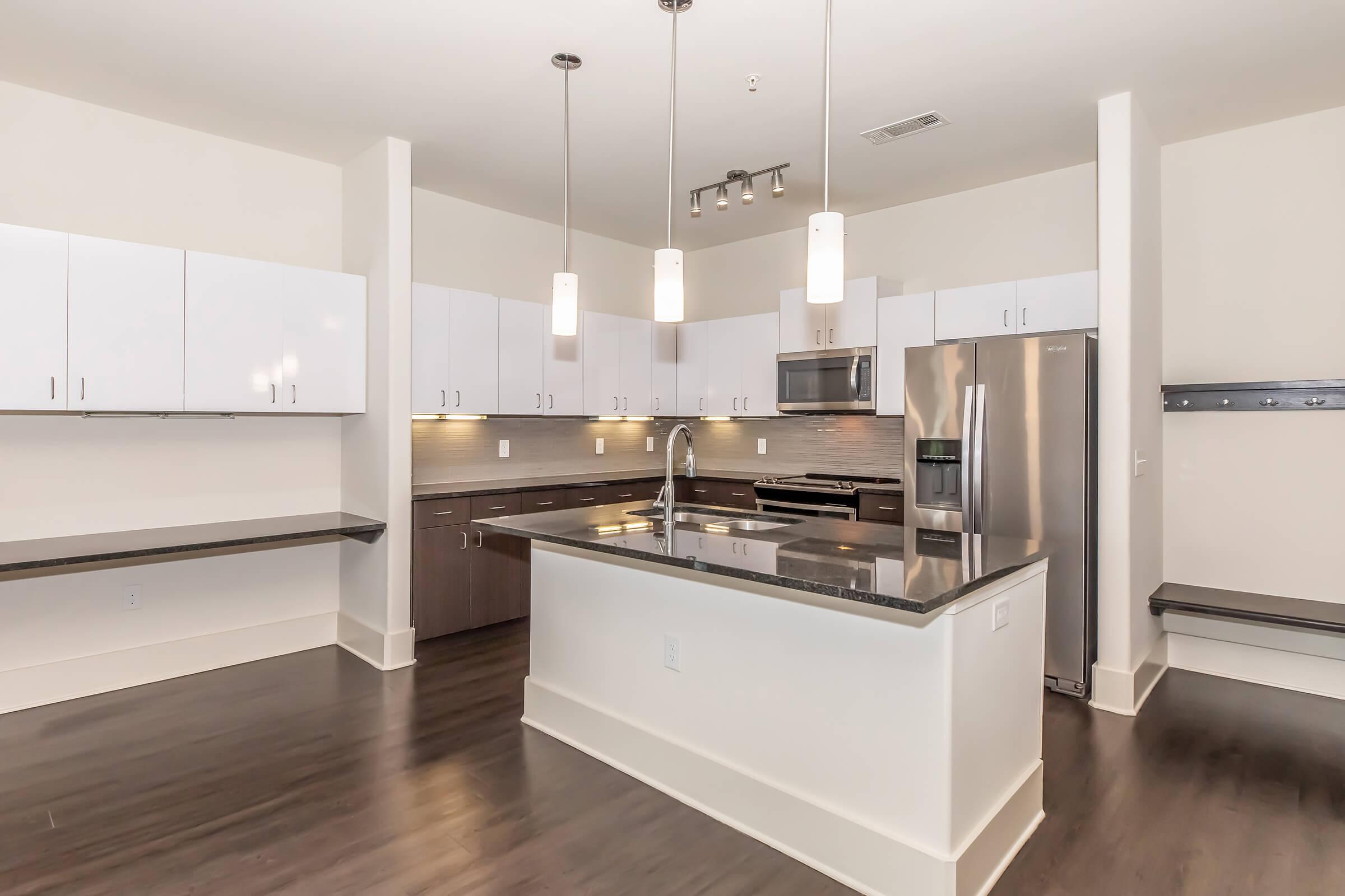 Modern kitchen featuring sleek cabinetry, stainless steel appliances, and a large island with dark countertops. The space has pendant lighting above the island, and an open layout with light-colored walls and dark wood flooring, creating a contemporary and inviting atmosphere.