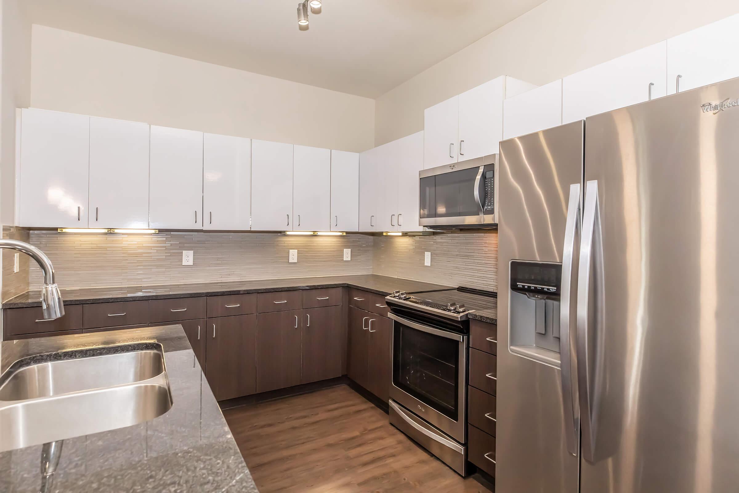 Modern kitchen featuring sleek stainless steel appliances, including a refrigerator and oven, with a built-in microwave. The cabinets are a mix of dark wood and glossy white finishes. A granite countertop surrounds a double sink, and light-colored tile backsplashes add a contemporary touch to the overall design.