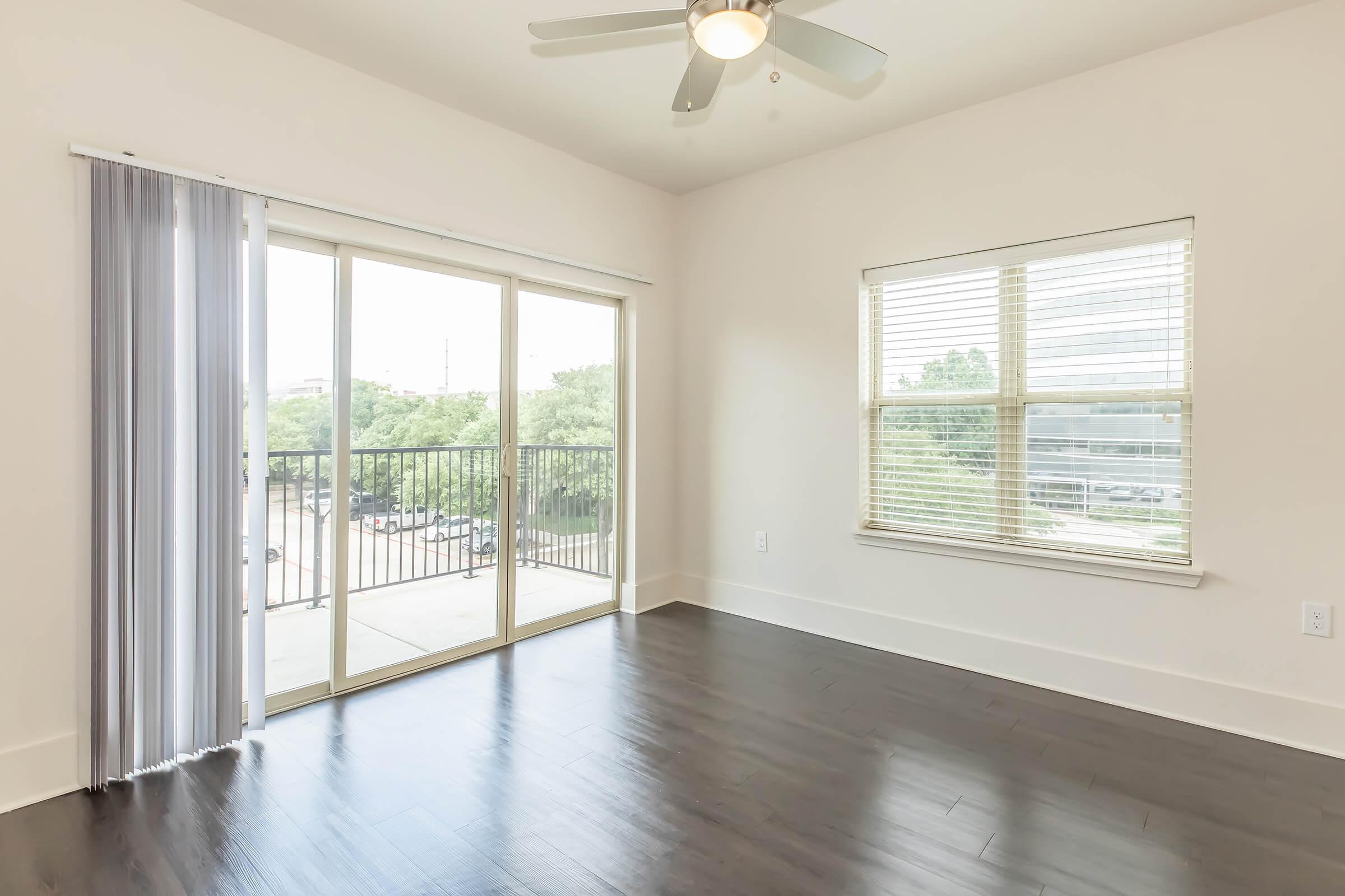 Bright and spacious room with hardwood floors, featuring a ceiling fan, large windows, and sliding glass doors leading to a balcony. Natural light fills the space, creating a welcoming atmosphere. There are white walls and a simple design, ideal for modern living.