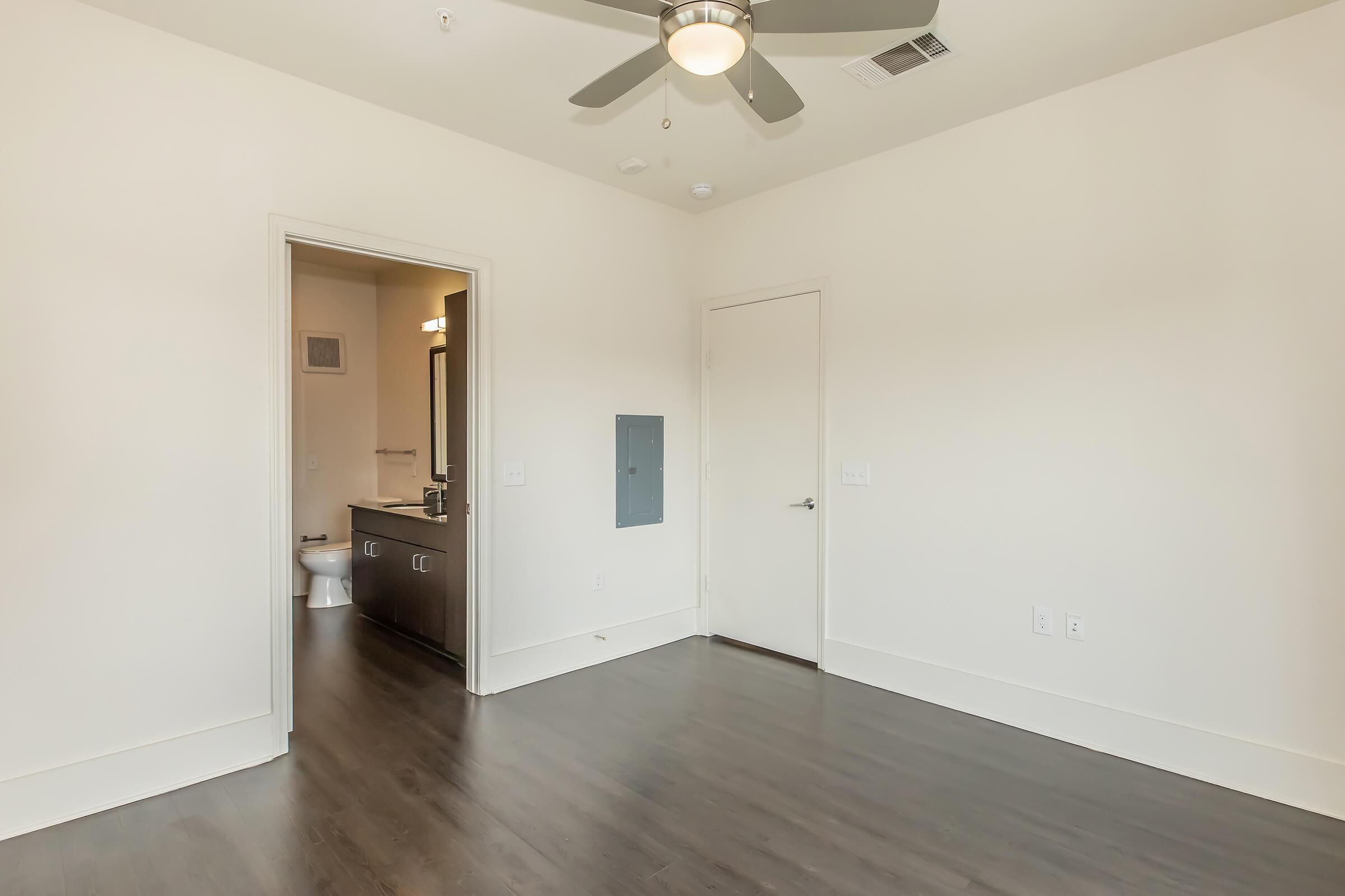 A spacious room with light-colored walls and a dark wood floor. A ceiling fan is mounted above. In the background, a doorway leads to a bathroom with a sink and toilet visible. The room has a modern, minimalistic design and ample natural light.