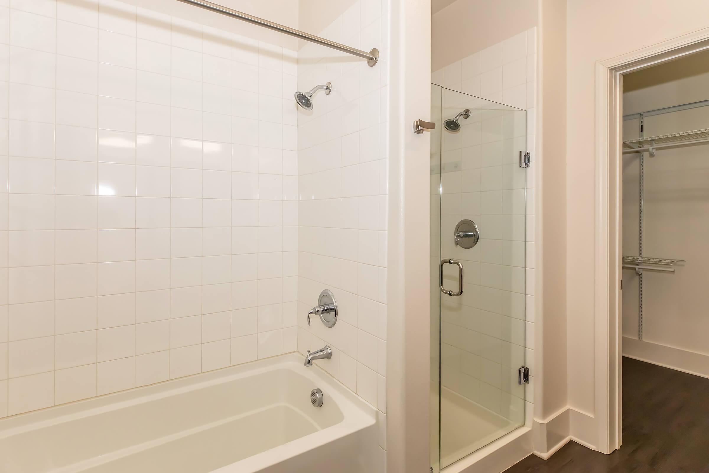 A modern bathroom featuring a white tiled wall with a bathtub on the left and a glass shower enclosure on the right. The shower has a circular silver showerhead and a wall-mounted control. A door on the right leads to a closet with a shelving unit. The flooring is dark, contrasting with the light tiles.