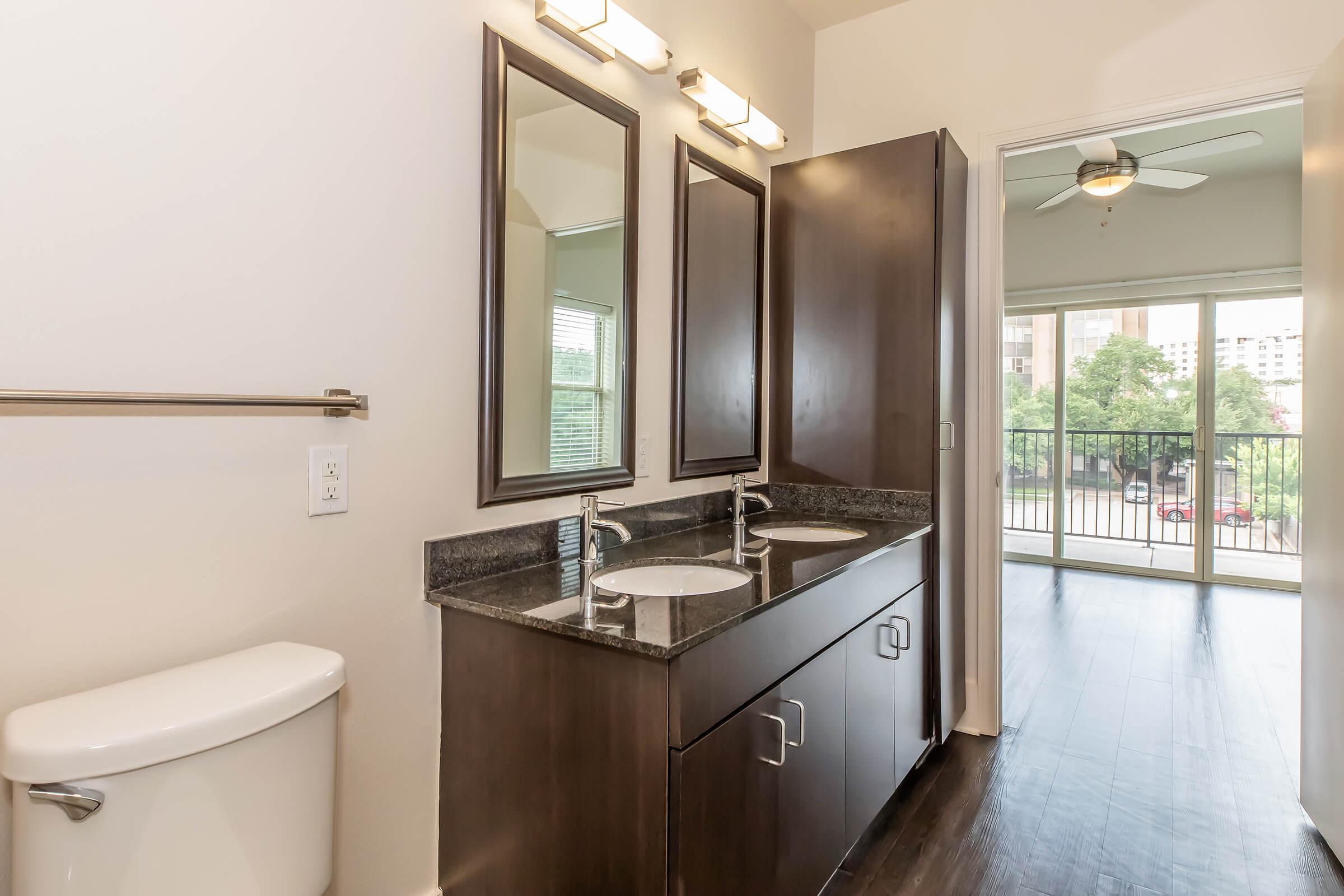 Modern bathroom with dual sinks and dark wood cabinetry. It features two mirrors, a toilet, and a large window with a view of the outdoors. The flooring is dark, enhancing the contemporary feel of the space.