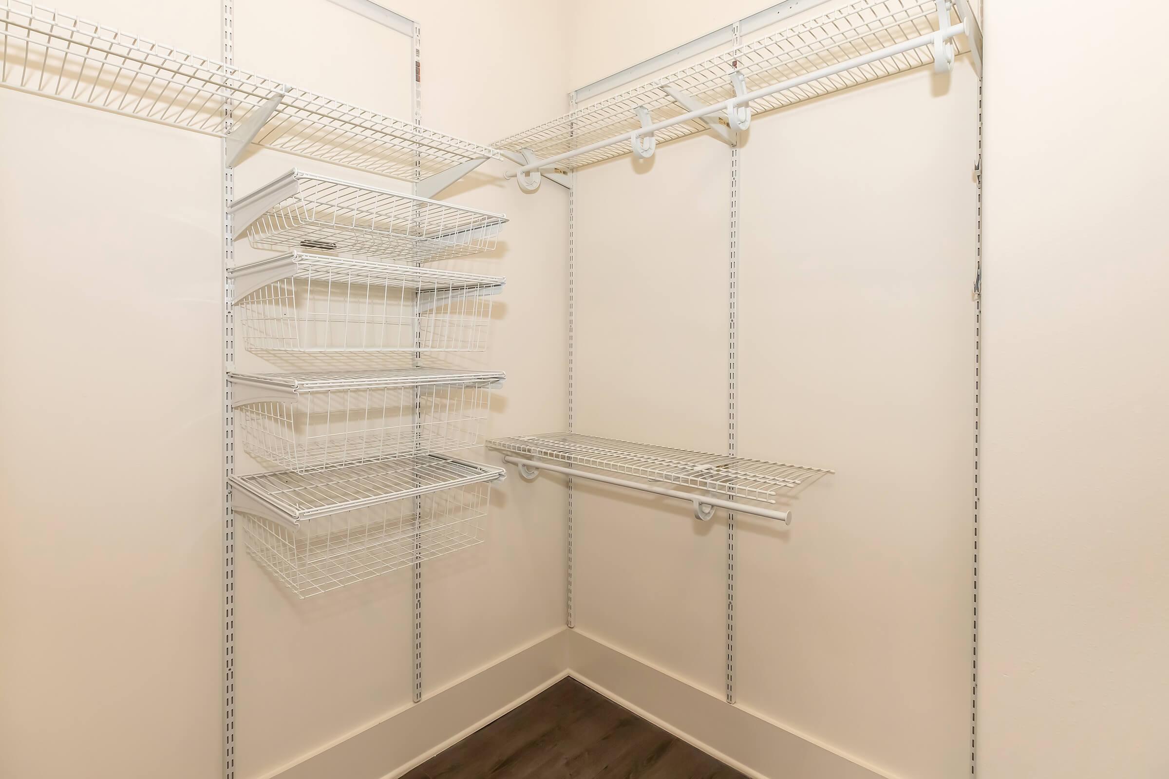 A spacious, empty closet featuring white wire shelving, including multiple levels of storage baskets and a flat shelf. The walls are a light color, and the floor is dark wood, creating a clean and organized appearance, ideal for storing various items.