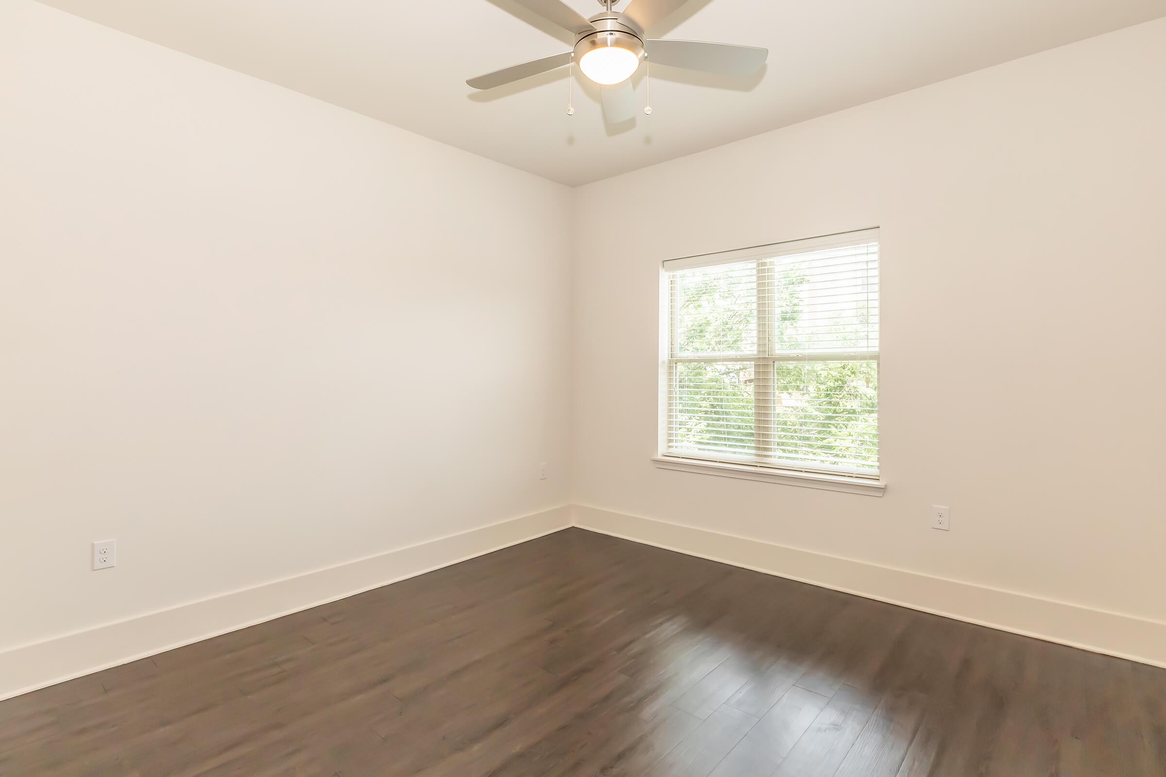 A clean, empty room with light-colored walls and dark hardwood flooring. A ceiling fan is mounted in the center, and a window with blinds allows natural light to enter, offering a view of greenery outside. The overall atmosphere is bright and inviting.