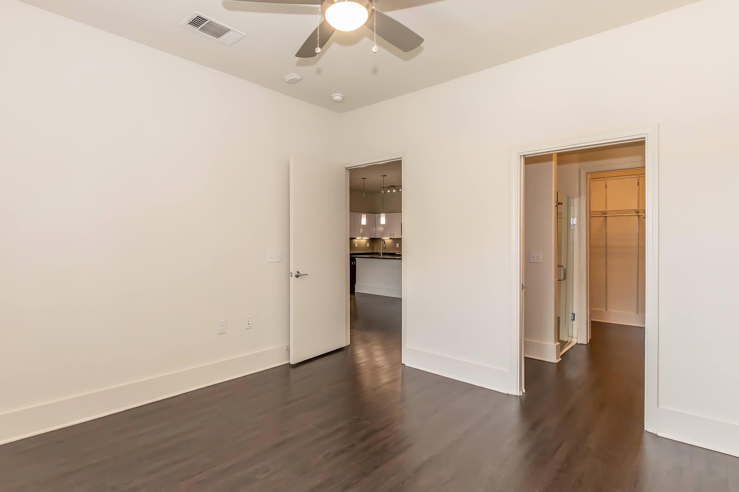 A spacious, empty room featuring dark hardwood floors, a ceiling fan, and white walls. There is a doorway leading to a bathroom on the right and an open passage to a kitchen area in the background. Natural light illuminates the space, highlighting its modern design.