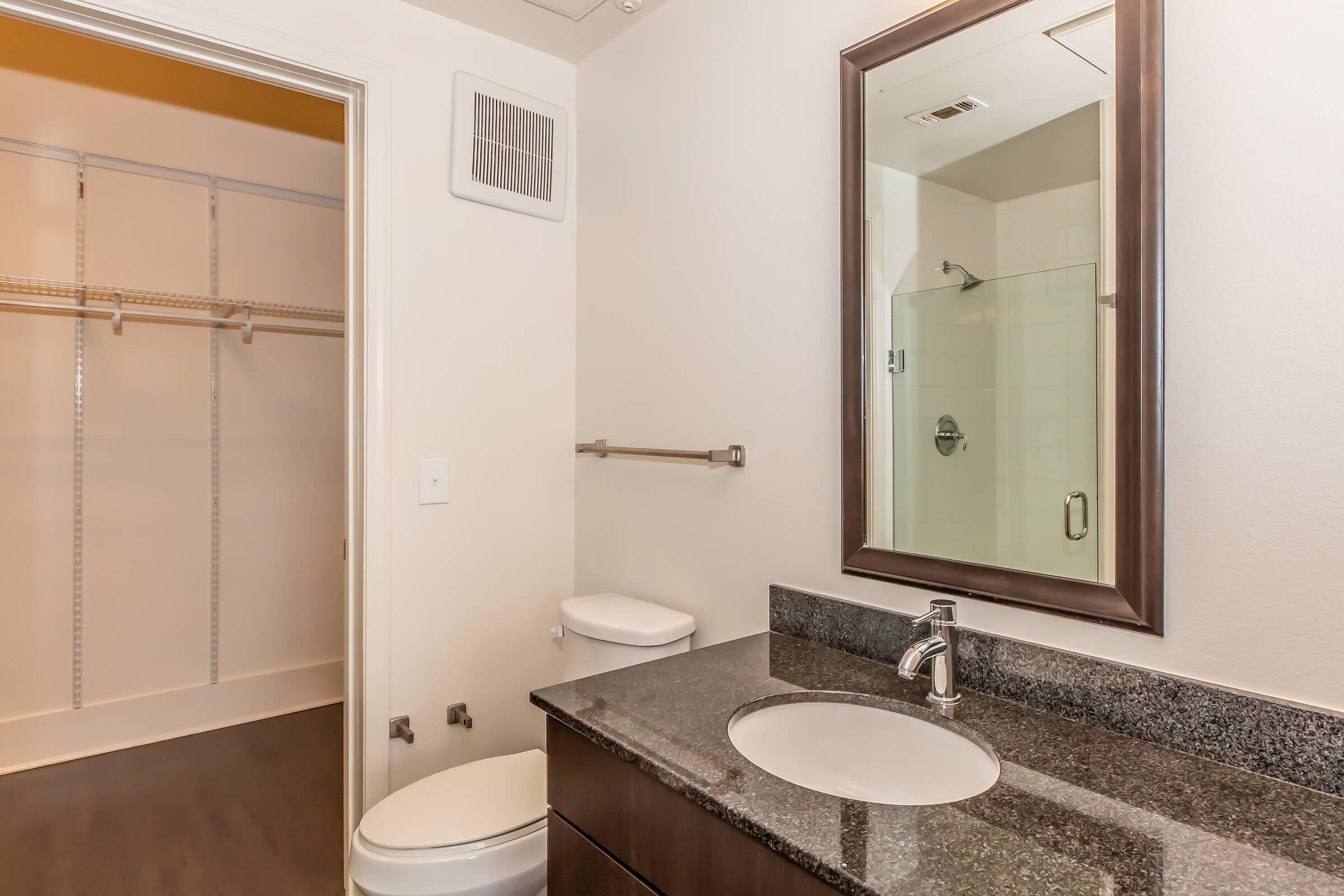 A modern bathroom featuring a granite countertop with a sink and mirror, a toilet, and a glass-enclosed shower. In the background, there is a spacious closet with wire shelving and a neutral-colored wall. The overall decor is simple and contemporary.