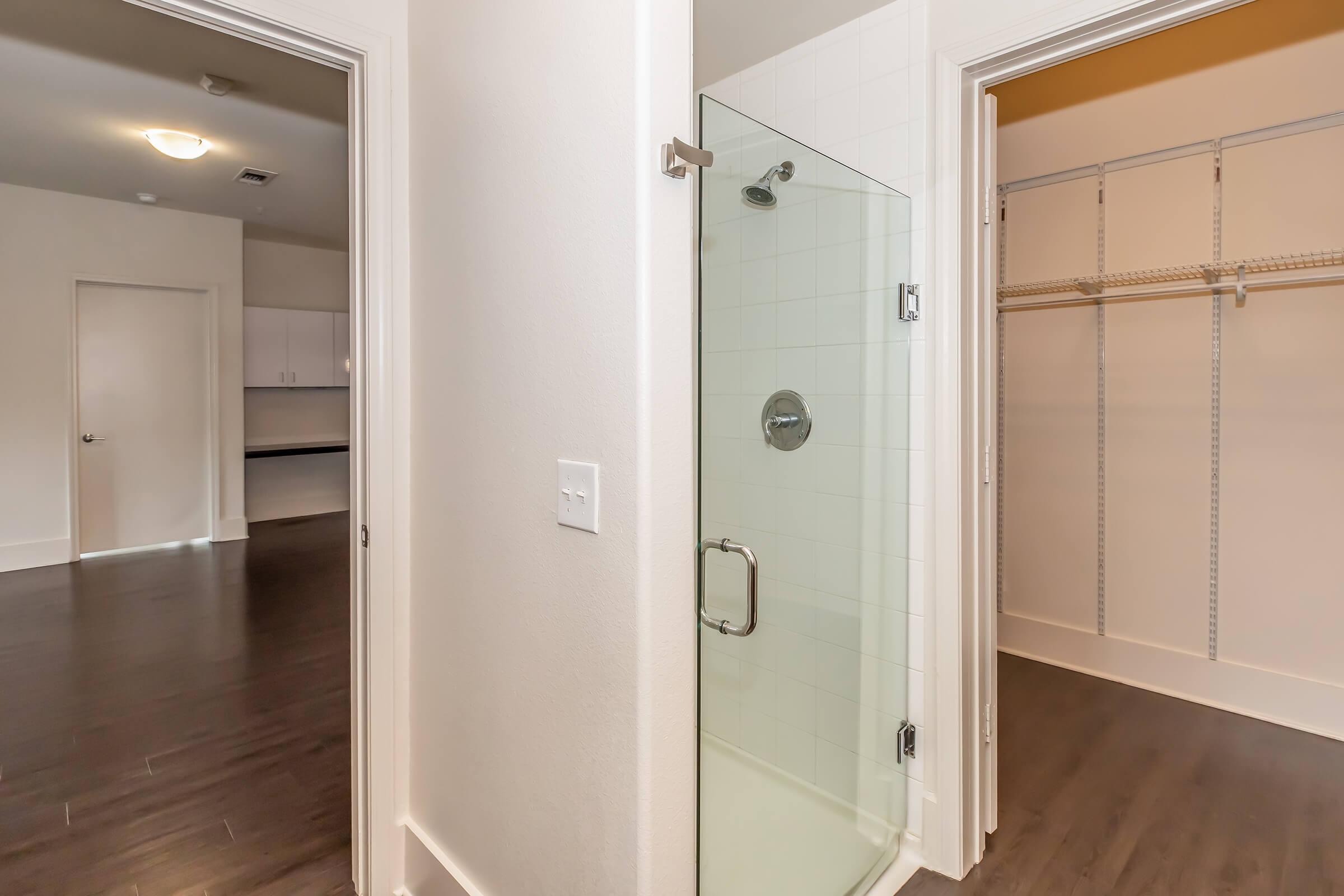 View of a bathroom corner featuring a glass shower enclosure, with a door leading to a spacious closet on the right. In the background, there is a glimpse of a clean kitchen area, showcasing modern design with hardwood flooring and bright lighting.