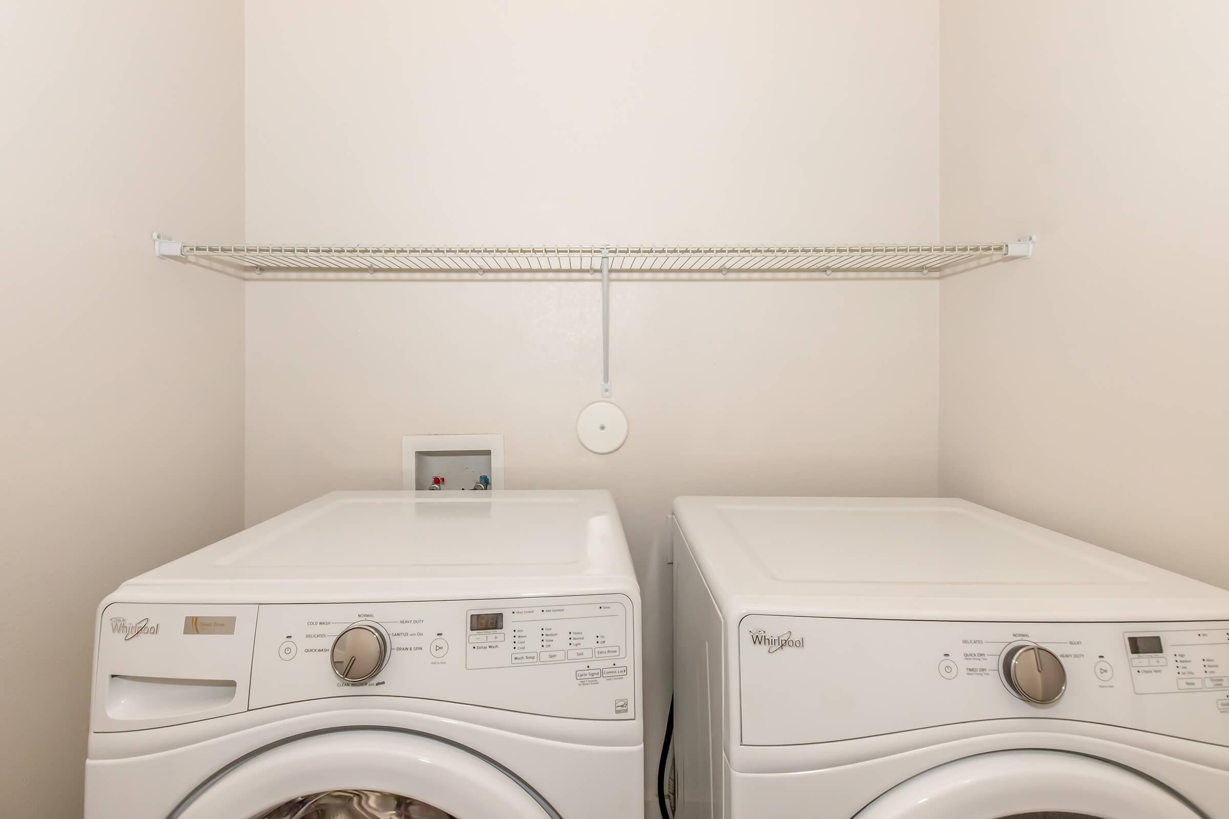 A clean laundry room featuring two white Whirlpool washer and dryer units side by side. Above them, there is a wire shelf for storage. The walls are light-colored, creating a bright and tidy atmosphere.