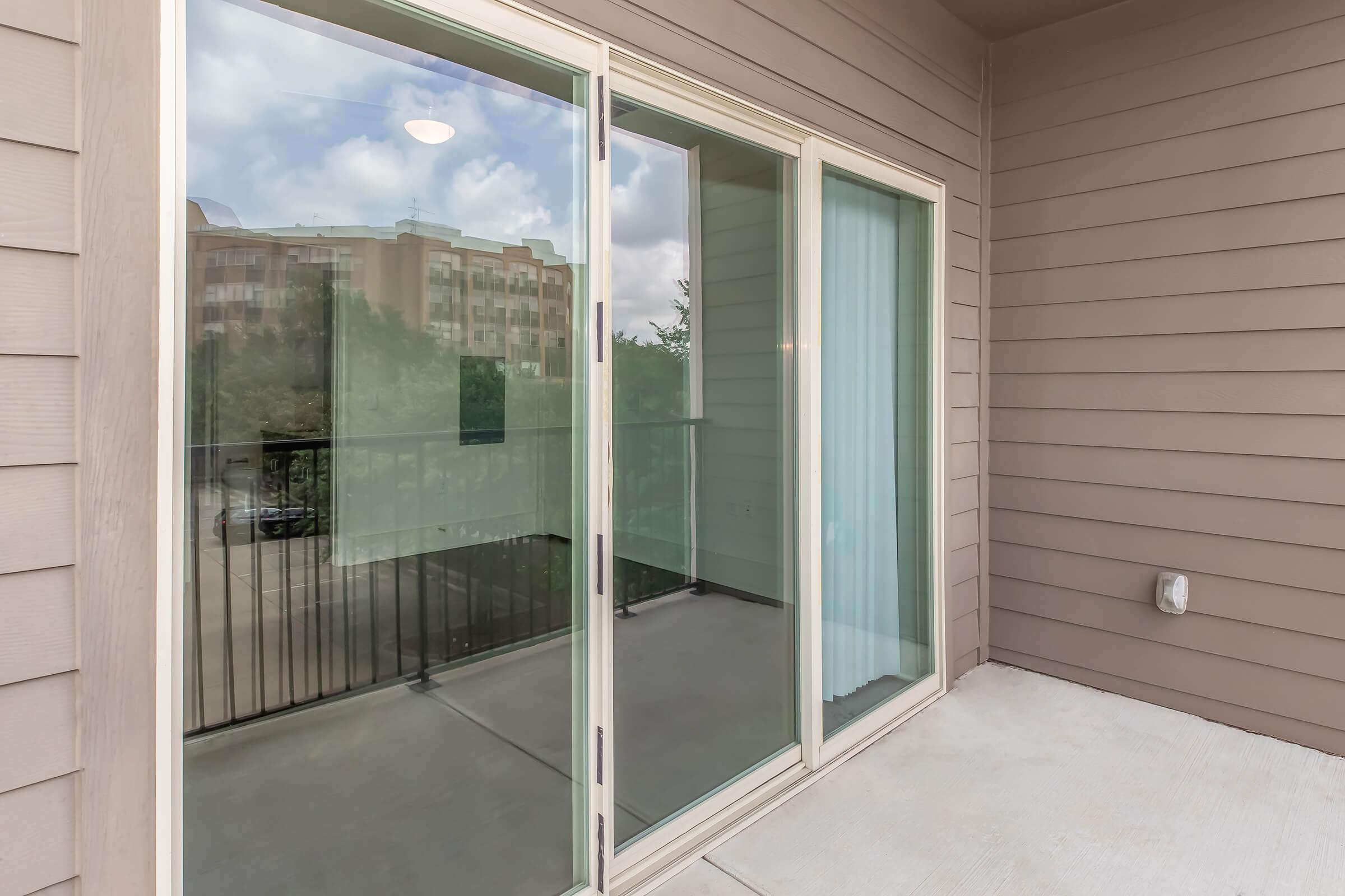 A view of a modern balcony with sliding glass doors, featuring a neutral-toned exterior wall and floor. The balcony is empty and offers a glimpse of the surrounding area, including trees and nearby buildings in the background. The overall atmosphere is bright with natural light.