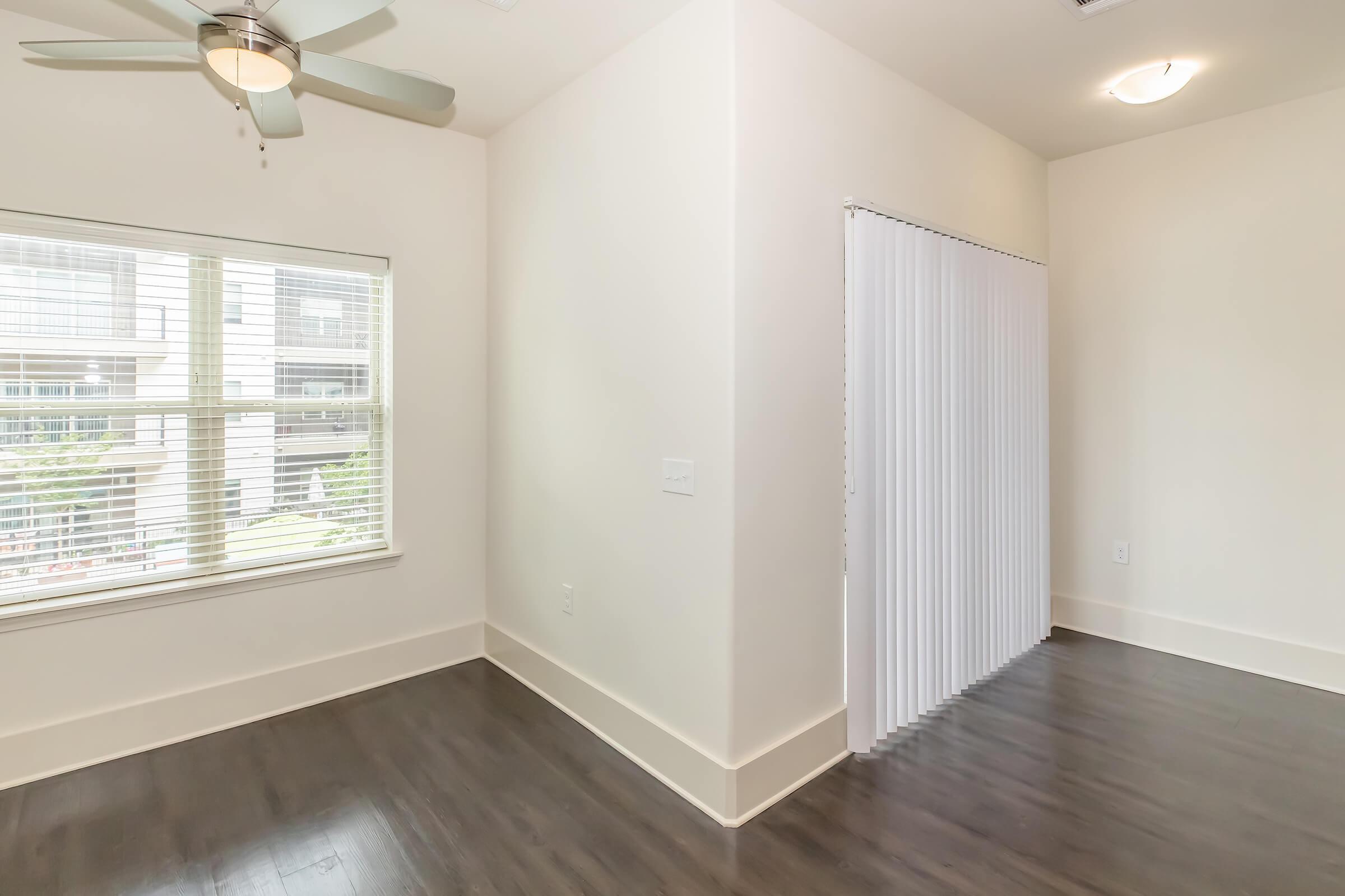 A bright, modern room featuring a ceiling fan and large window with blinds. The floor is dark wood, and a white partition is visible, separating the space. Natural light fills the room, creating an inviting atmosphere.