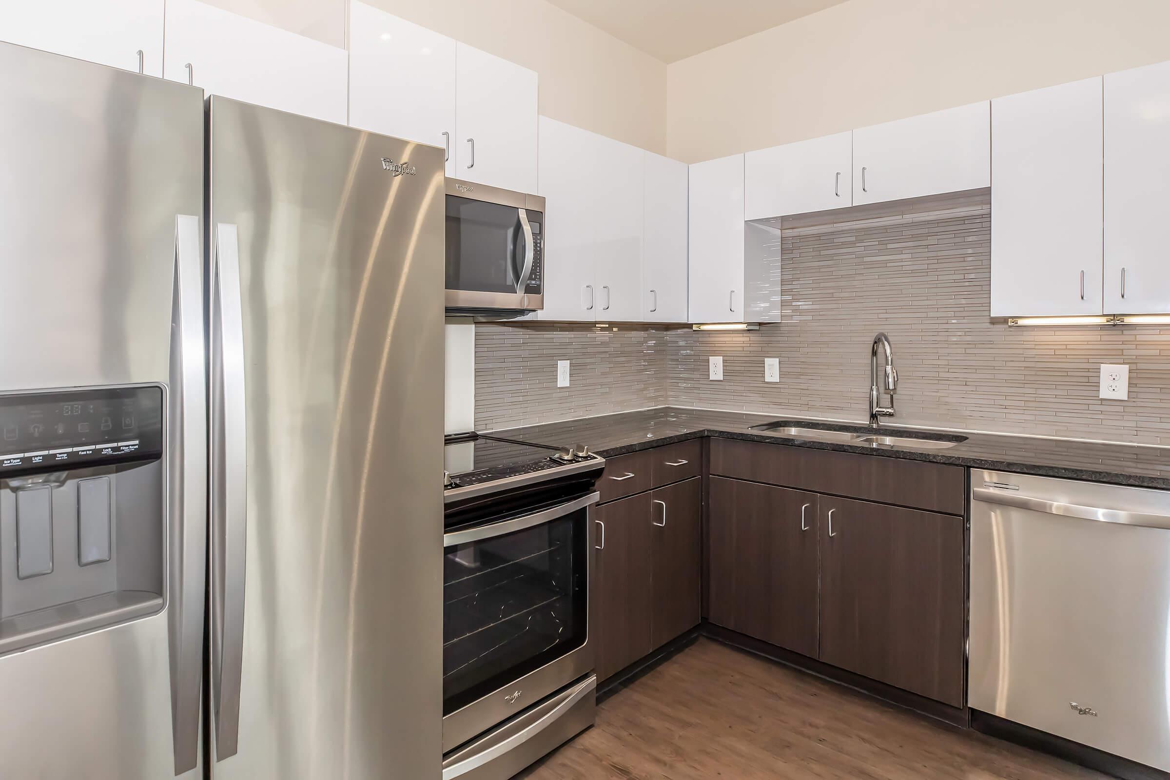 Modern kitchen featuring stainless steel appliances, including a refrigerator and oven, along with a microwave. Cabinets are a mix of white and dark wood. The countertop is dark granite, and the backsplash consists of light-colored tiles. Well-lit and spacious design suitable for cooking and dining.