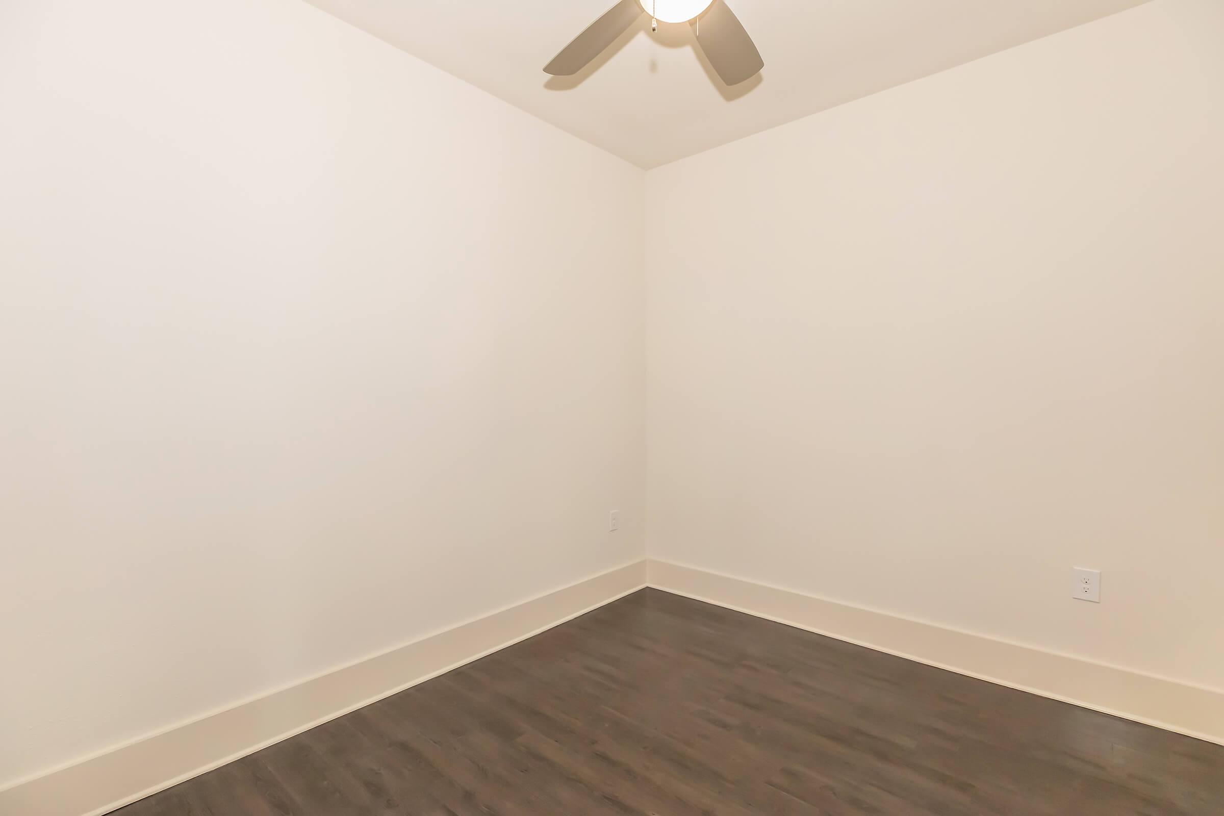 Empty room with light-colored walls and dark wooden flooring. A ceiling fan is visible, and there are no furniture or decorations present. The overall space appears minimalist and ready for customization.